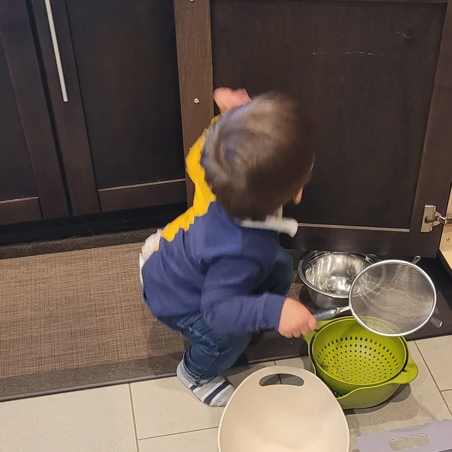 Toddler crouching at an open low kitchen cabinet, pulling out a colander, a strainer and bowls