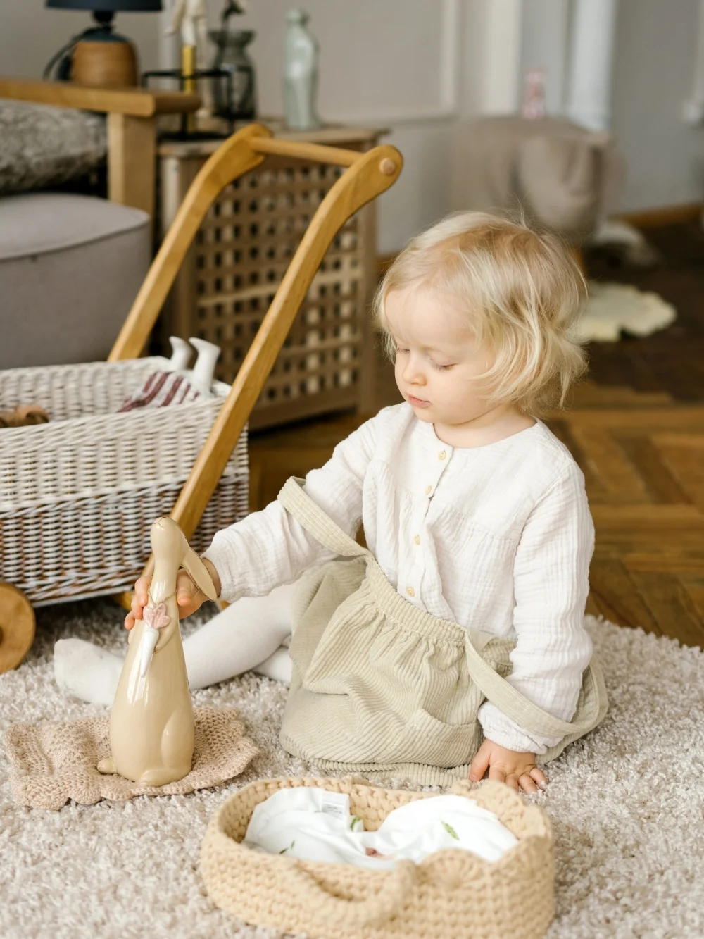 A toddler sits on a rug beside a small basket, playing with a toy bunny