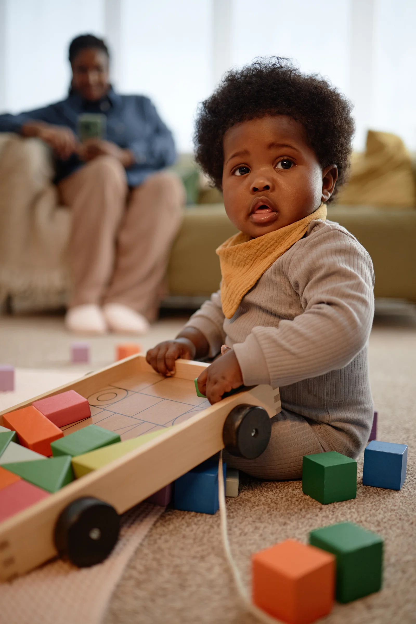 Toddler stacking wooden blocks on a rug while a parent sits nearby, watching