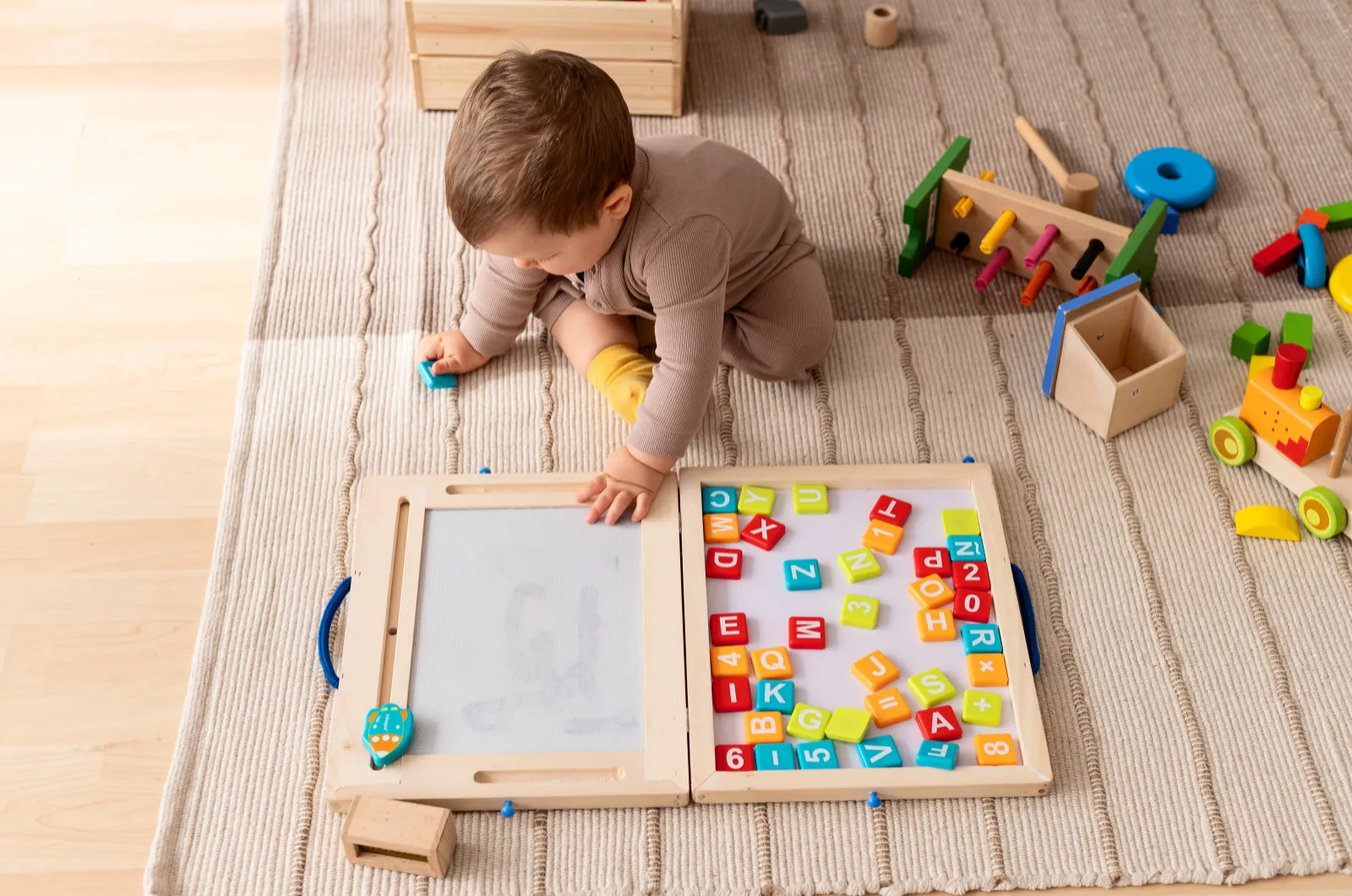 A toddler sits on a woven rug, absorbed in a wooden magnetic letter board, with more wooden toys nearby