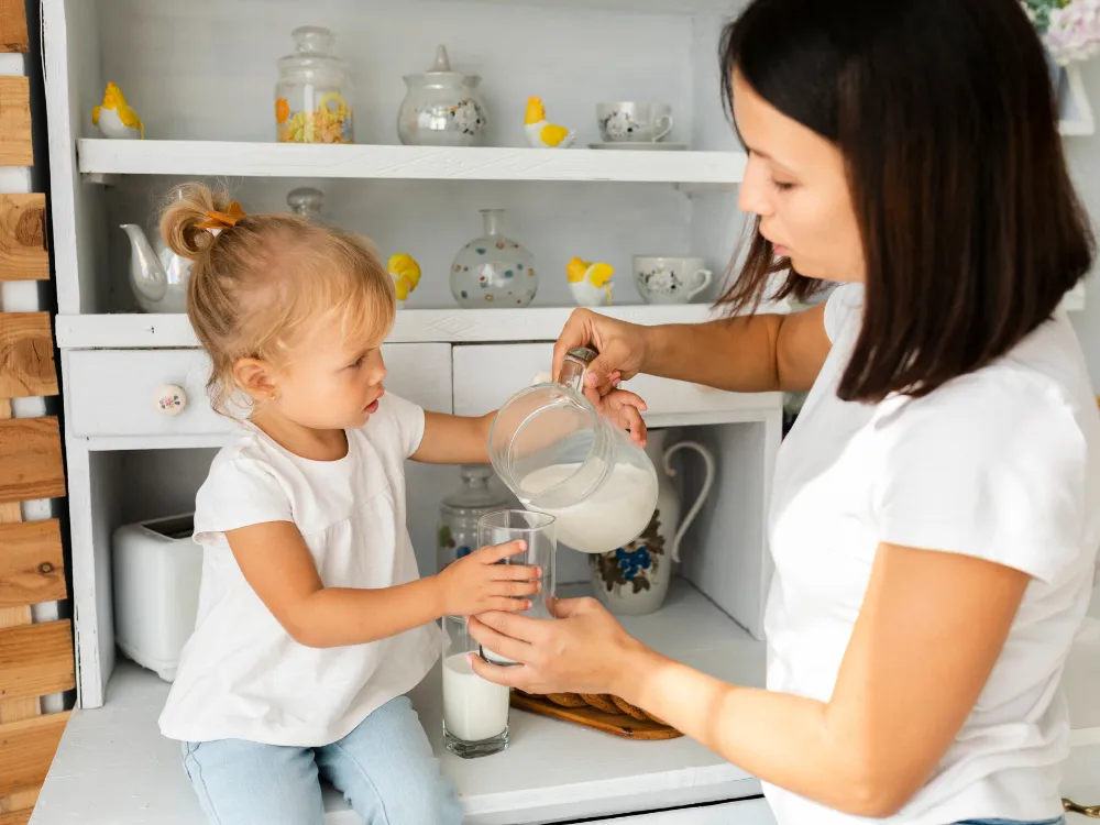 A toddler holds a glass steady while a grown-up helps pour milk from a small jug