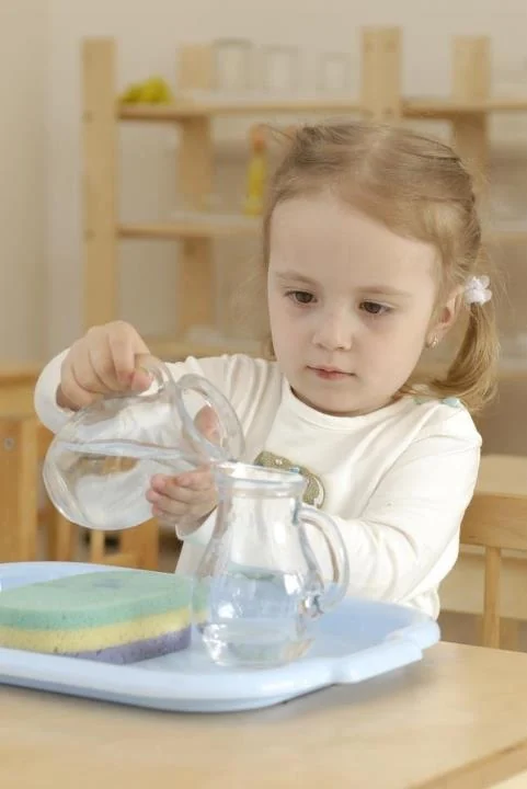 Toddler girl pouring water from one small glass pitcher into another, with a sponge on the tray beside them
