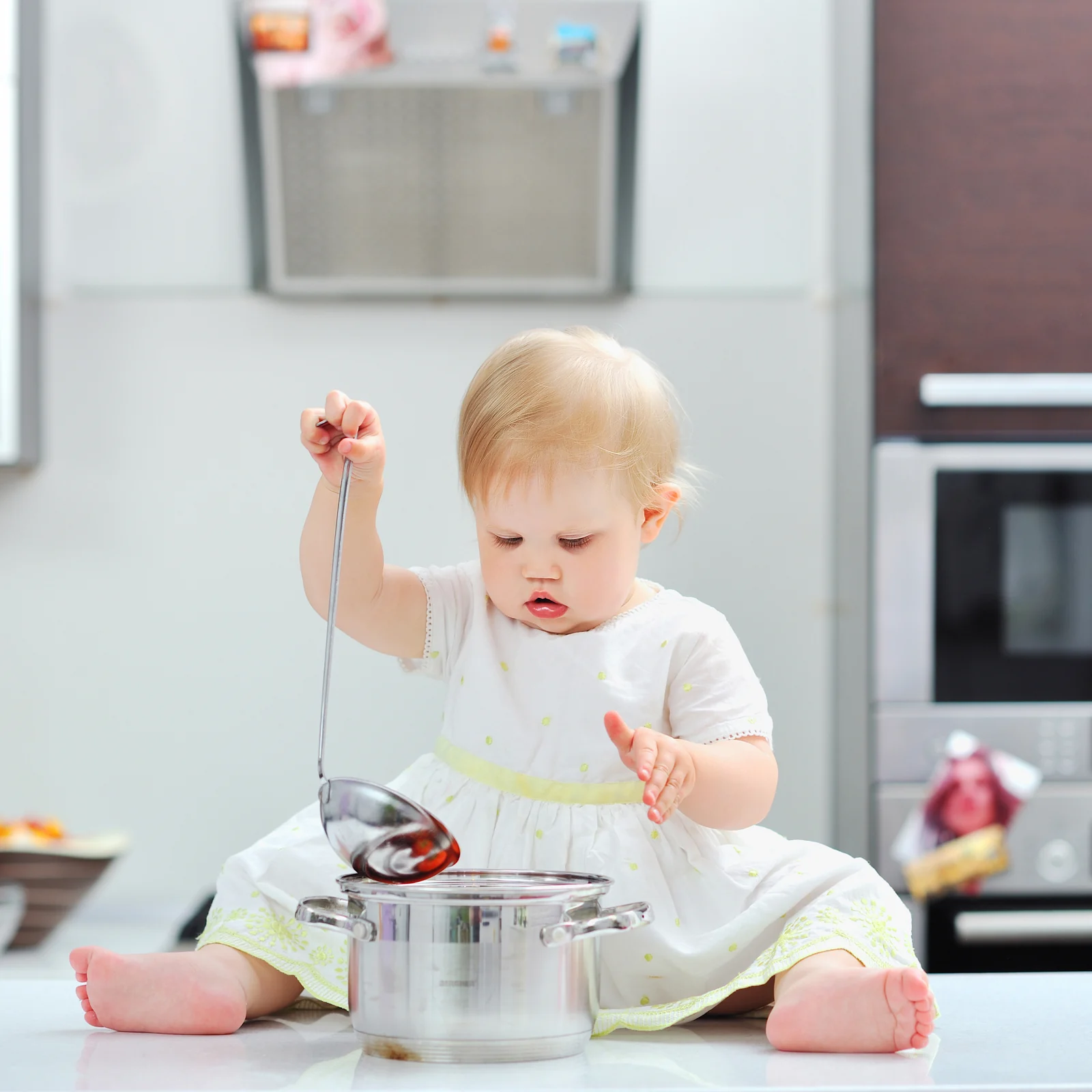 Toddler sitting on the kitchen floor with an empty metal pot, pretending to cook with a ladle