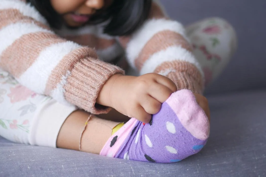 Toddler sitting on the floor, concentrating hard on pulling on their own sock
