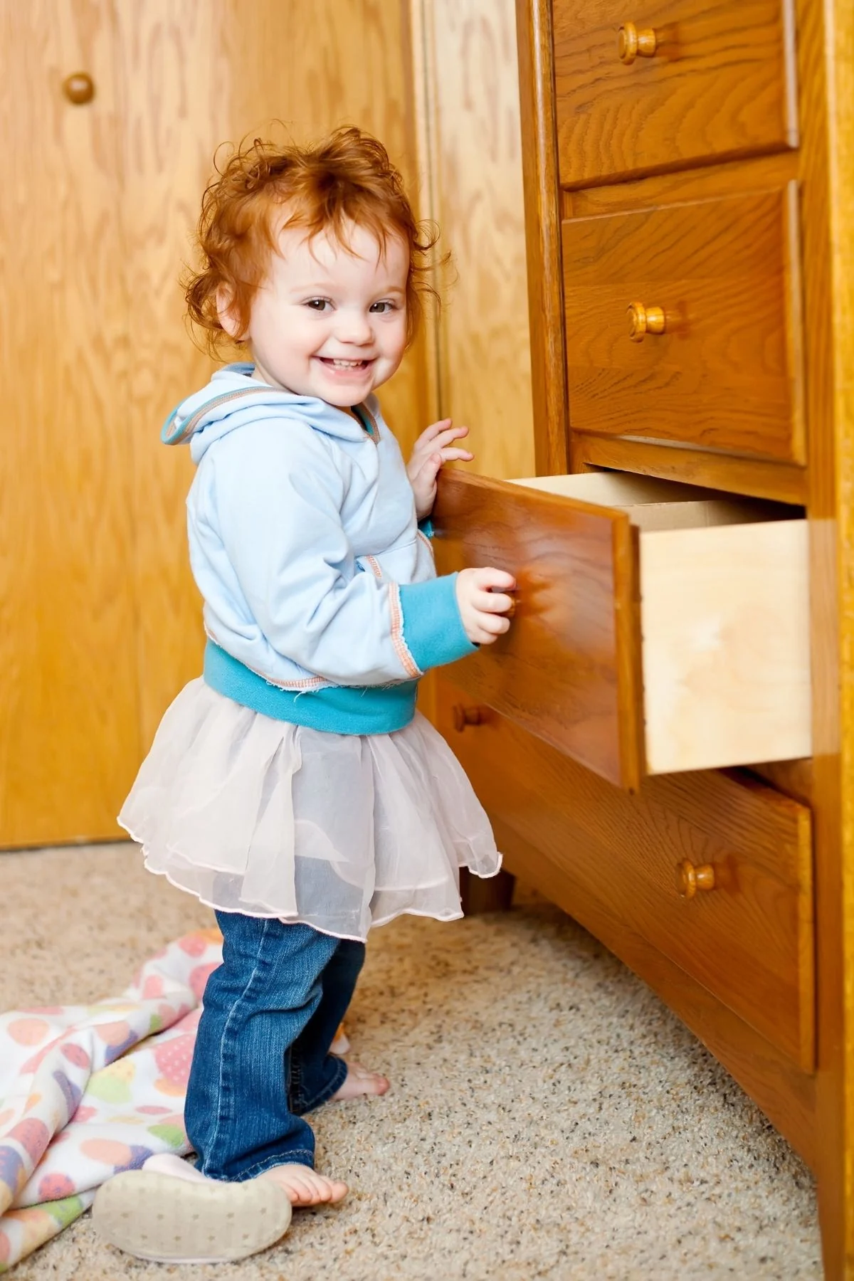 A smiling toddler in a blue hoodie pulls open a drawer of a wooden dresser