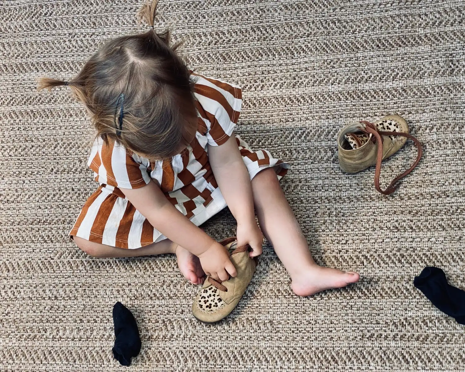 Toddler sitting on a woven rug pulling a soft shoe onto her foot, with two socks beside her