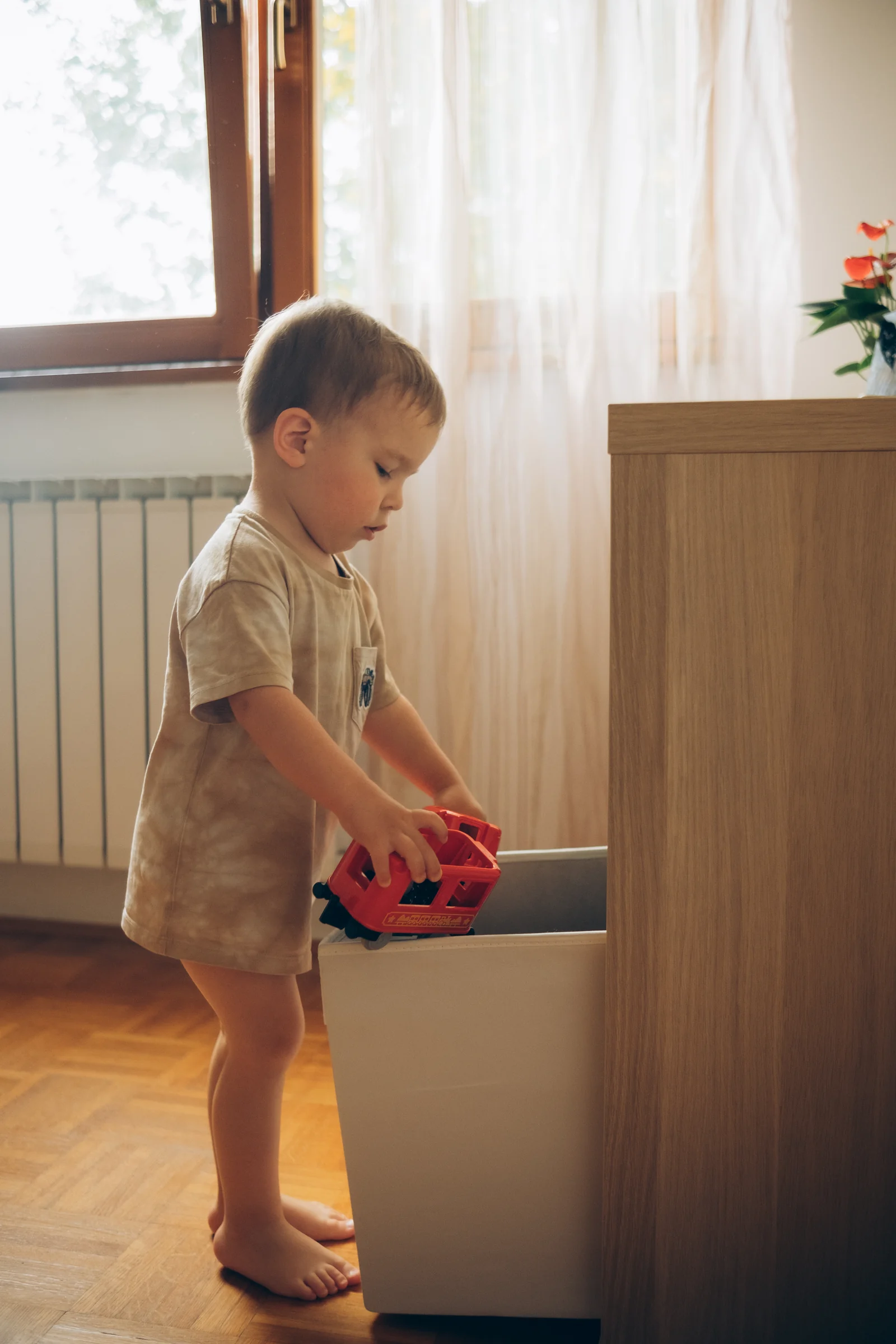 A toddler lowers a small red toy car into a white bin beside a wooden cabinet