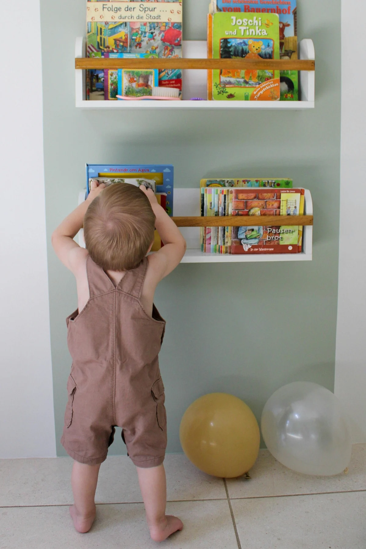 A toddler in brown overalls reaches up to a wall-mounted ledge shelf with picture books standing cover-out