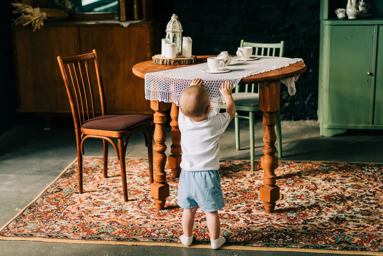 Toddler seen from behind on a patterned rug, reaching up to a table set with teacups, candles and a lace runner