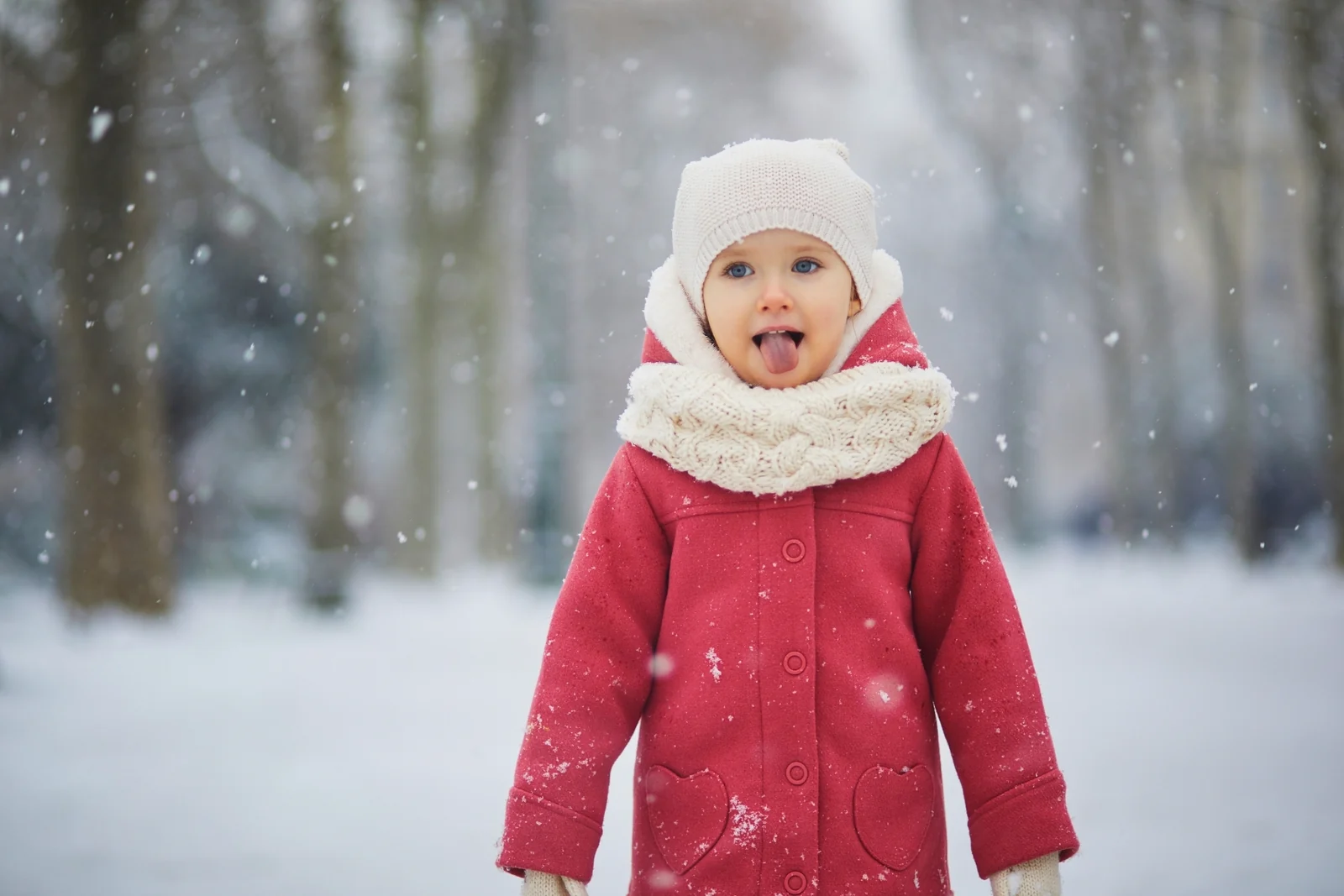 Toddler in a red coat, white knit hat and cream scarf standing outdoors in falling snow with tongue out to catch flakes