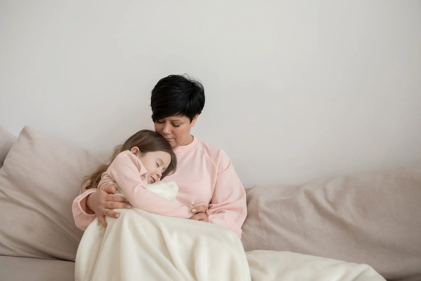 Young child in a pink sweatshirt dozing against a parent's chest on a couch, a cream blanket over their laps