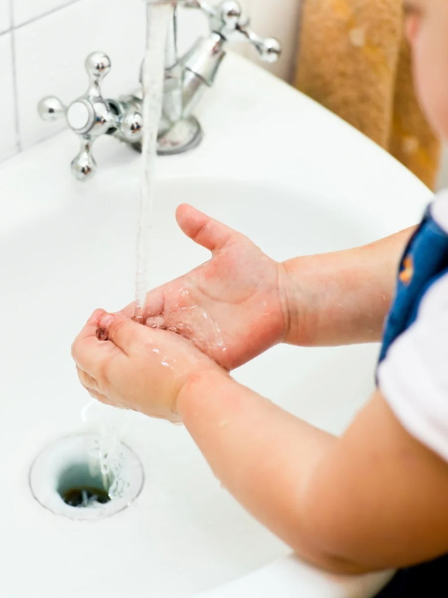 A toddler rinses both hands under a running faucet in a white sink