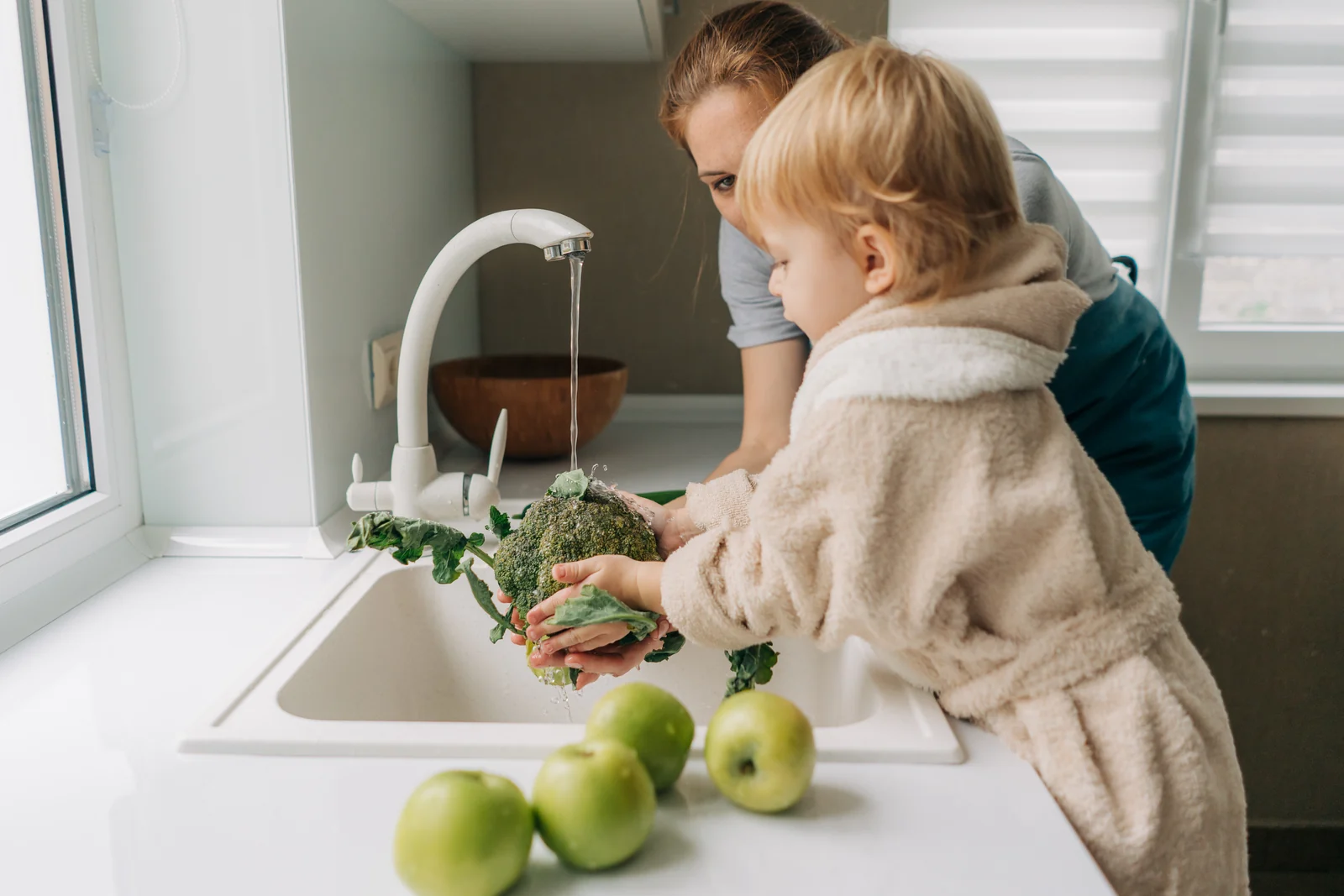 Toddler in a fluffy beige robe rinsing a head of broccoli under the kitchen tap while a parent leans in beside them