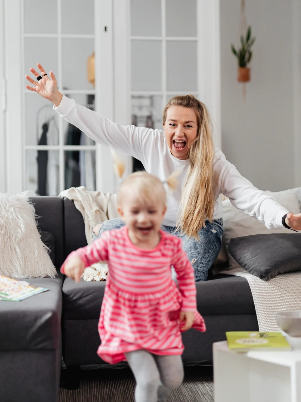 Laughing toddler in a pink striped dress running across a living room while an adult on the couch cheers with open arms