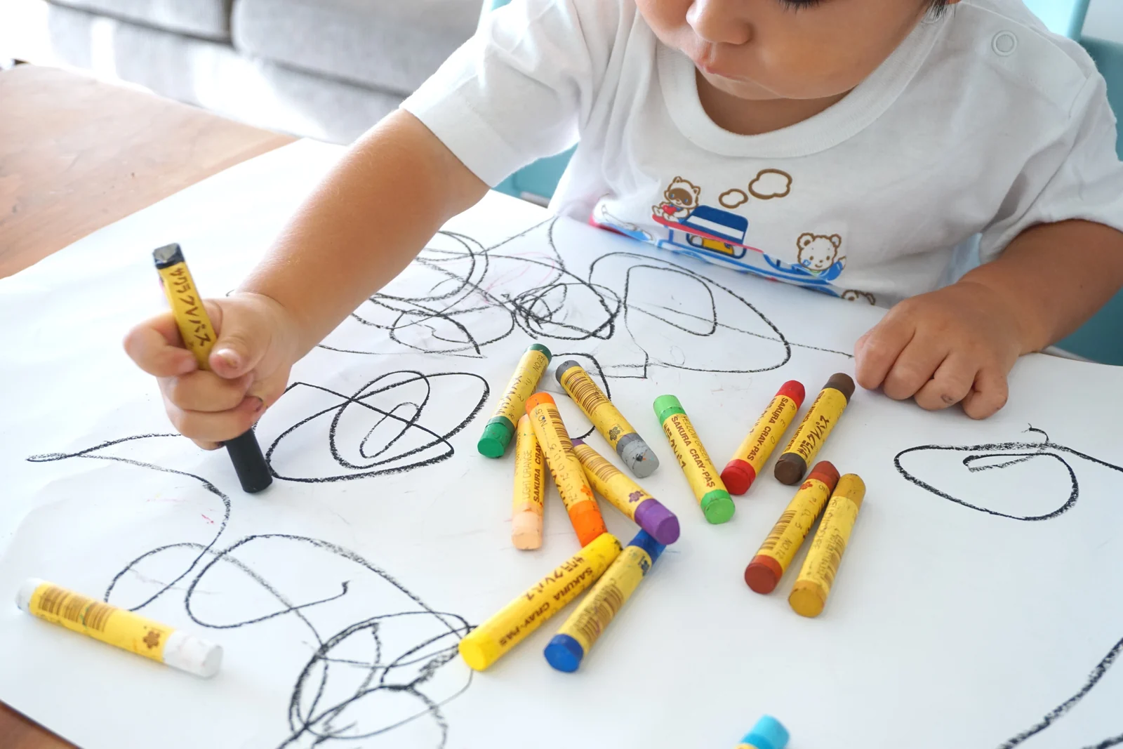 Toddler scribbling black loops on a big sheet of white paper with chunky wax crayons scattered around