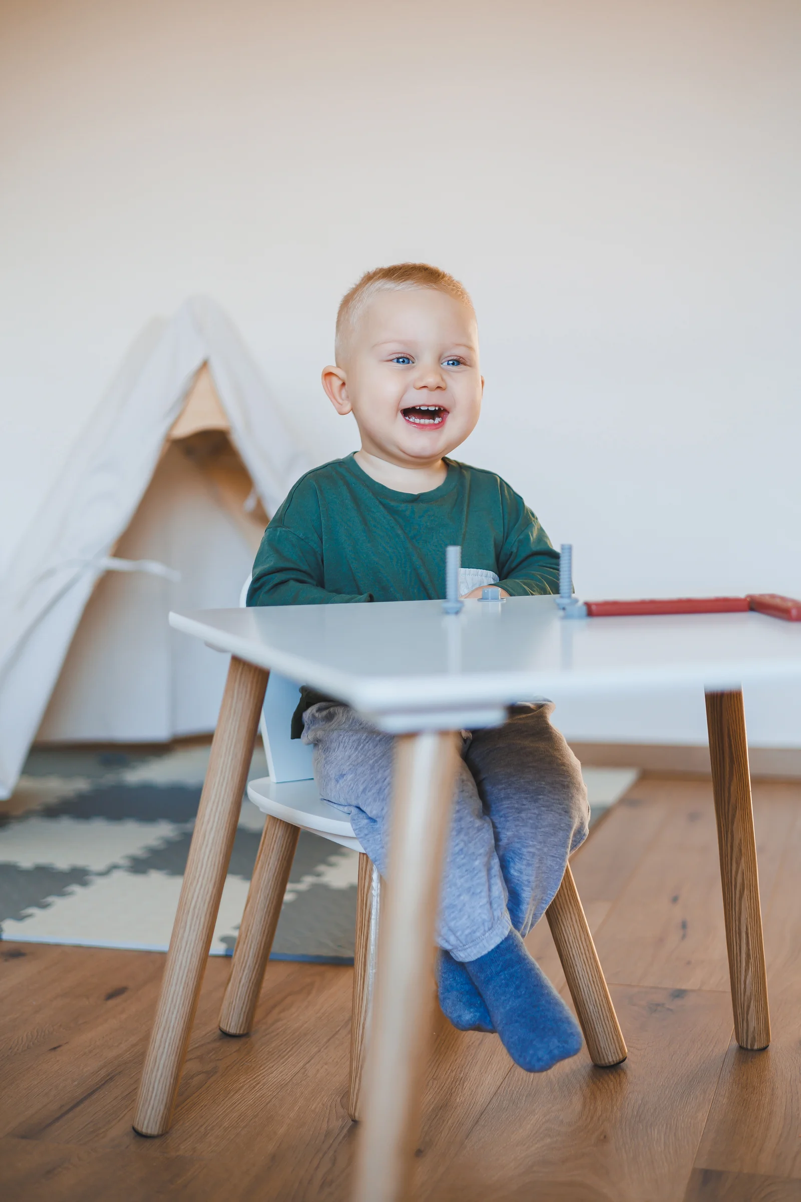 A smiling toddler sits on a child-sized chair at a small white table