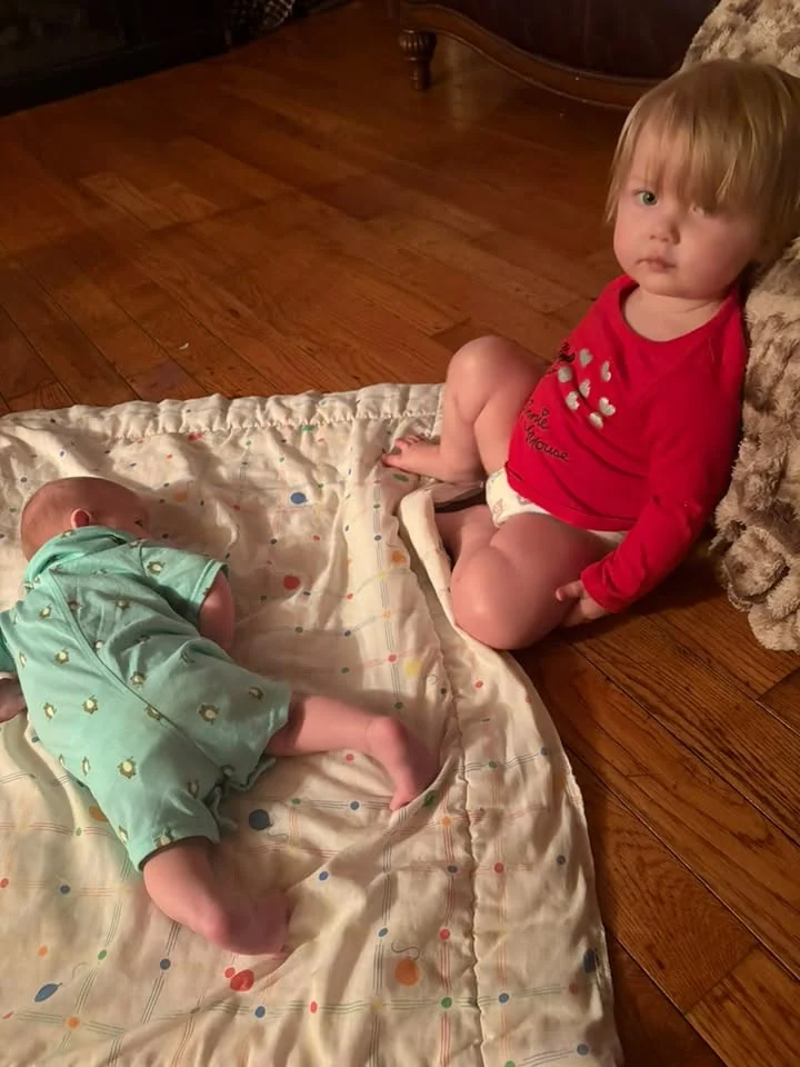 Toddler in a red top sitting on a wood floor beside a baby lying on a patterned quilt