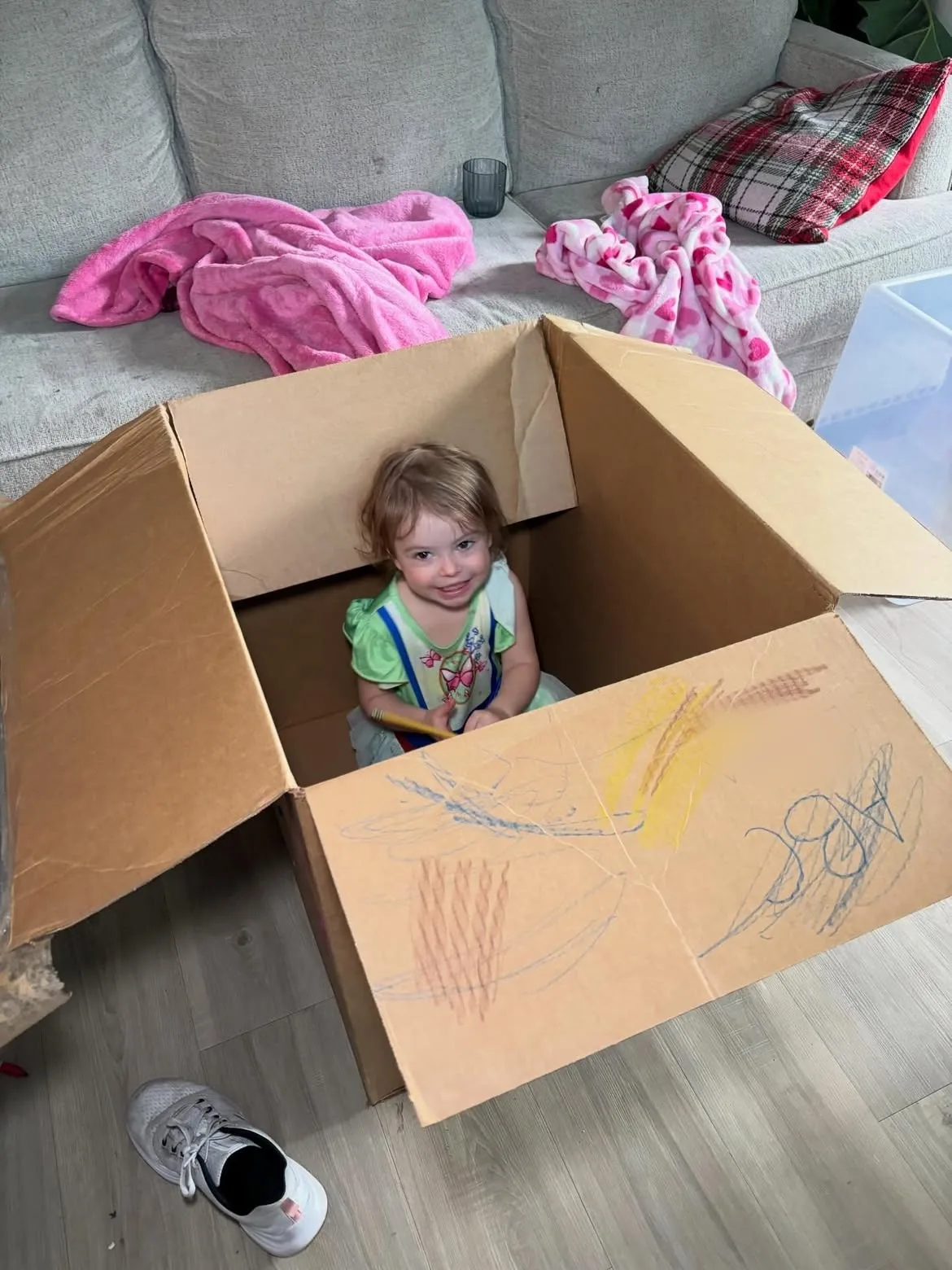 Smiling toddler sitting inside a large open cardboard box with crayon scribbles on its flap, a couch behind