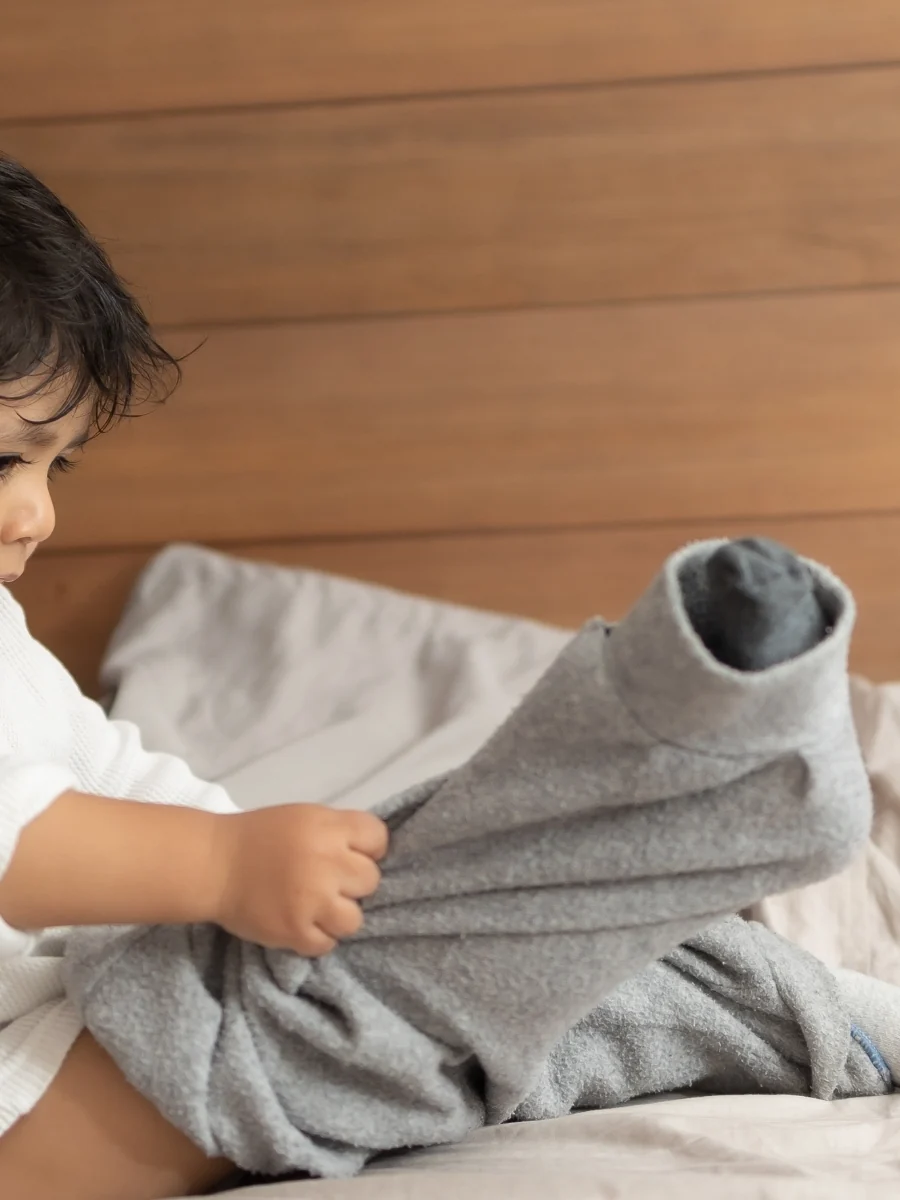 Toddler sitting on a bed pulling a pair of loose gray pants onto one leg