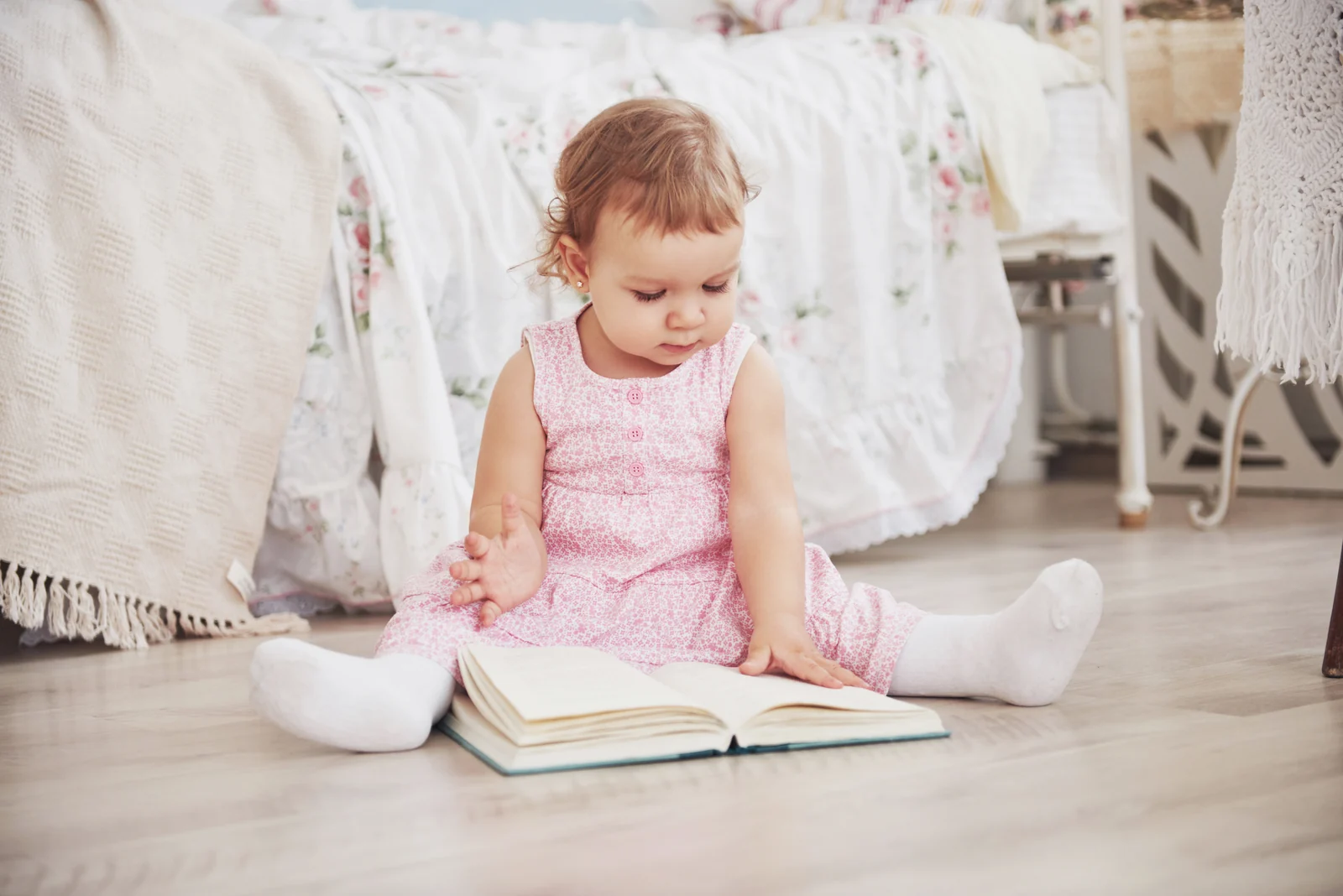 A toddler in a pink dress sits on a wooden floor with a large open book in front of her