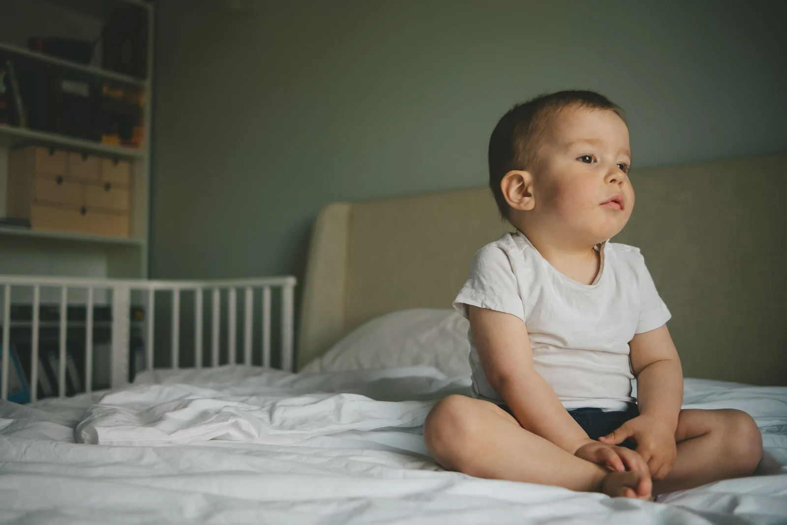 A toddler sitting up on a bed with white sheets, looking out across the room in soft light