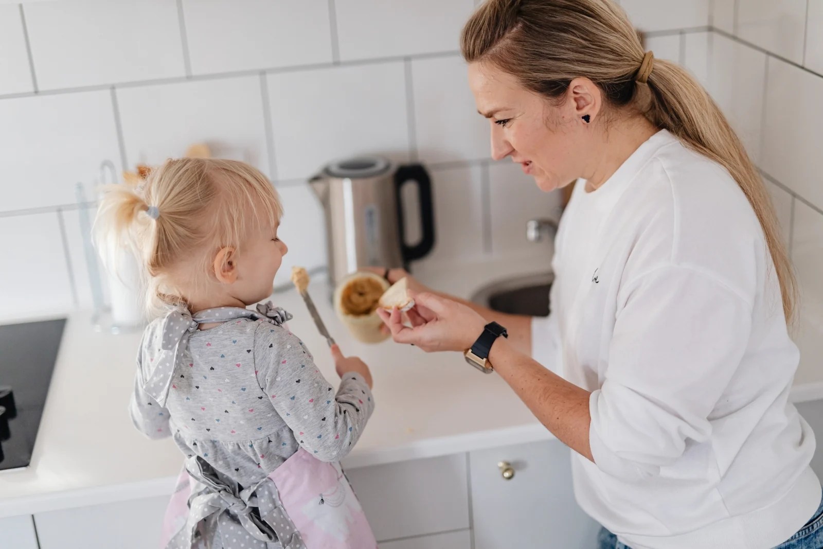 Toddler in an apron holding a knife with spread on it while a parent holds a slice of bread at the kitchen counter