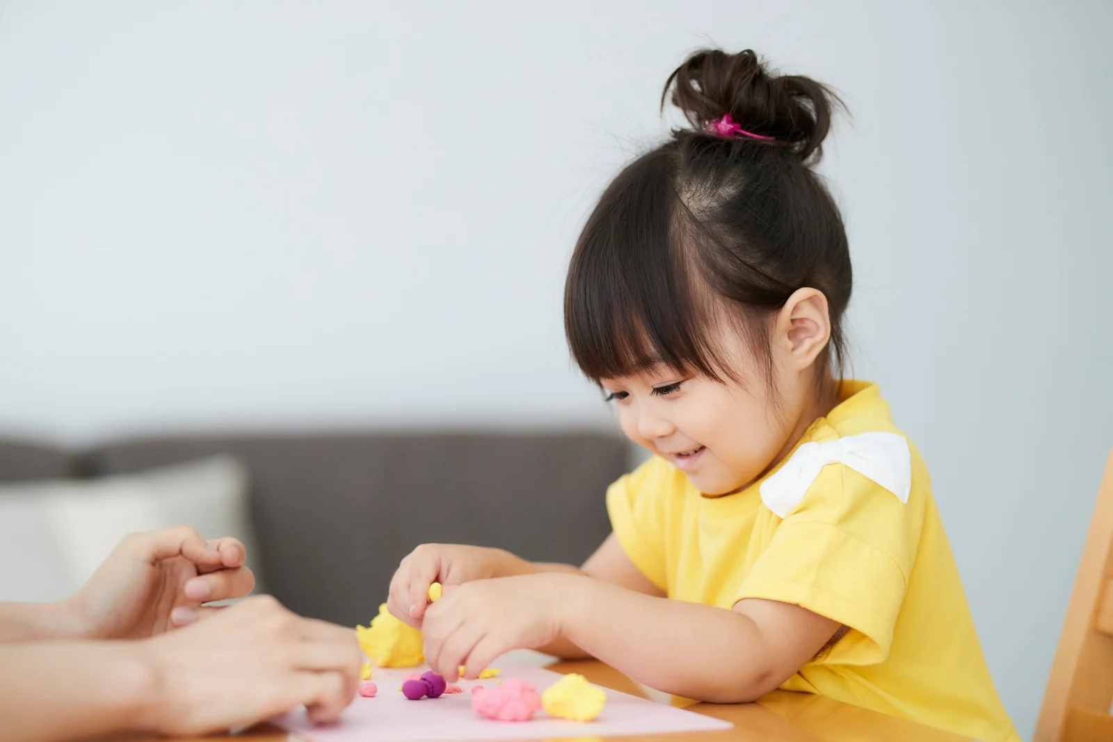 Child shaping small pieces of play dough at a table while an adult's hands rest nearby