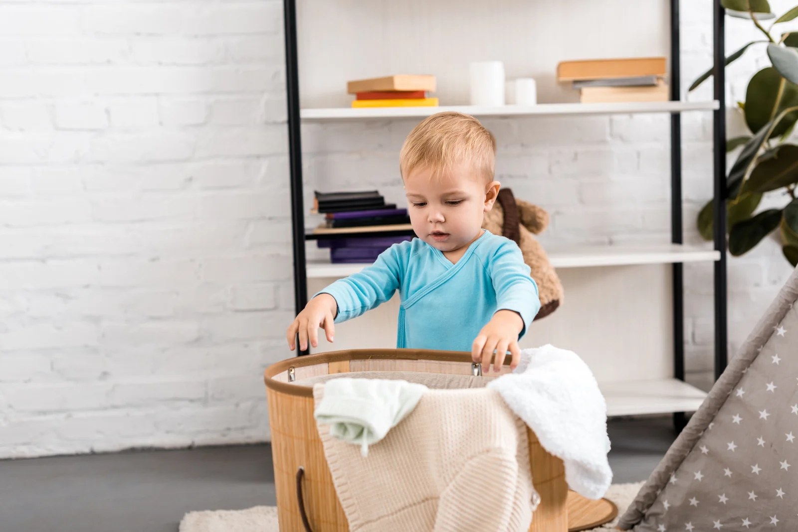 Toddler in a light blue top standing at a woven laundry basket piled with soft clothes, hands on the rim