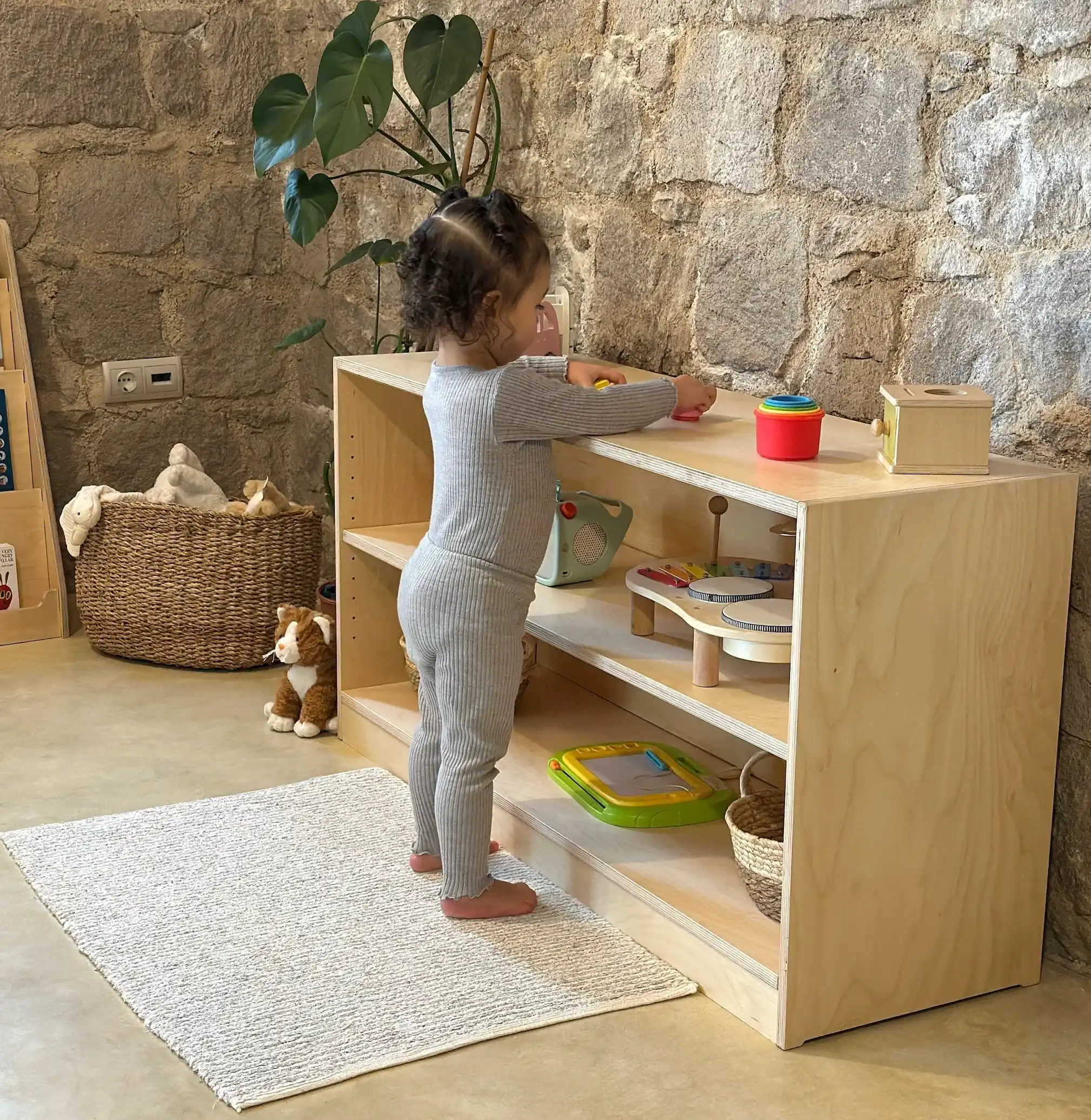 A toddler in a gray ribbed outfit stands at a low light-wood shelf, placing a toy on top, with a few toys and baskets below