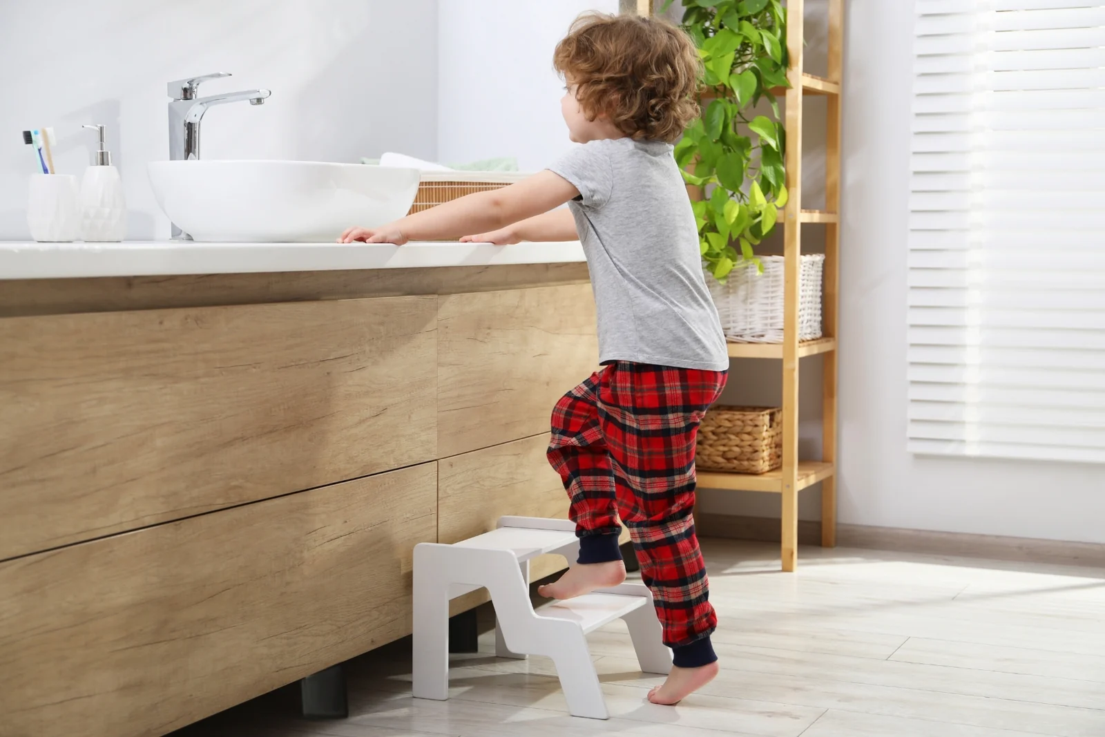 A toddler in red plaid pajama pants stepping onto a white two-step stool at a bathroom sink