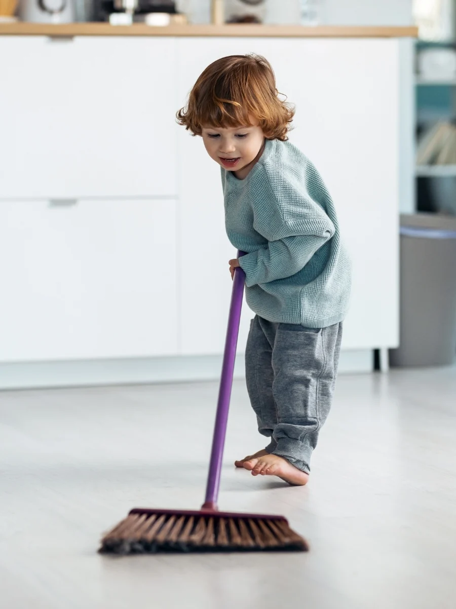 A toddler in a gray sweater pushes a small broom across a white kitchen floor