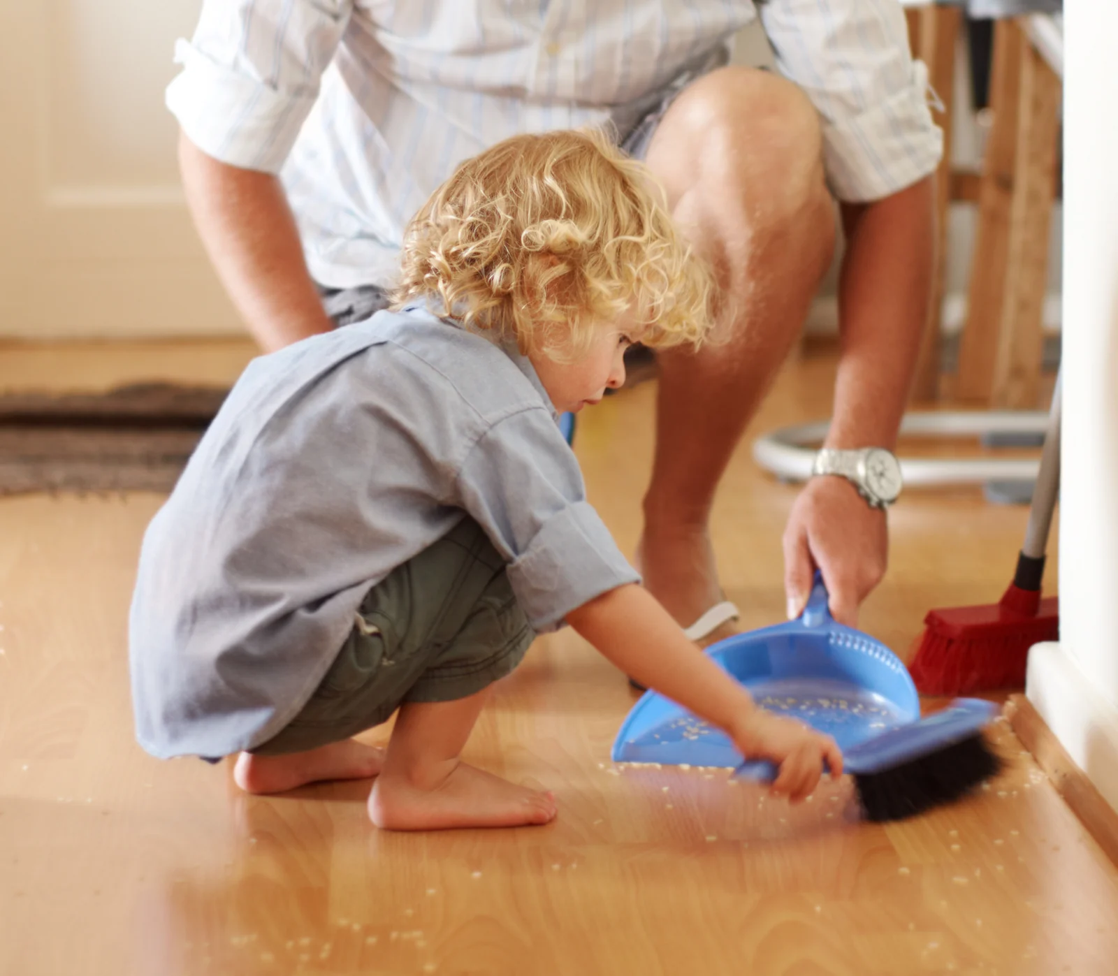 Toddler sweeping crumbs into a blue dustpan on a wood floor while a parent crouches beside them