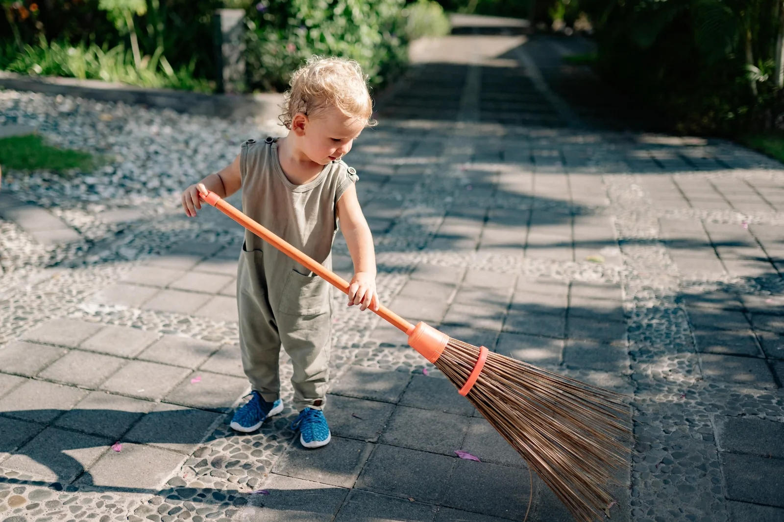 Toddler in an olive jumpsuit sweeping a stone-paved garden path with a large broom