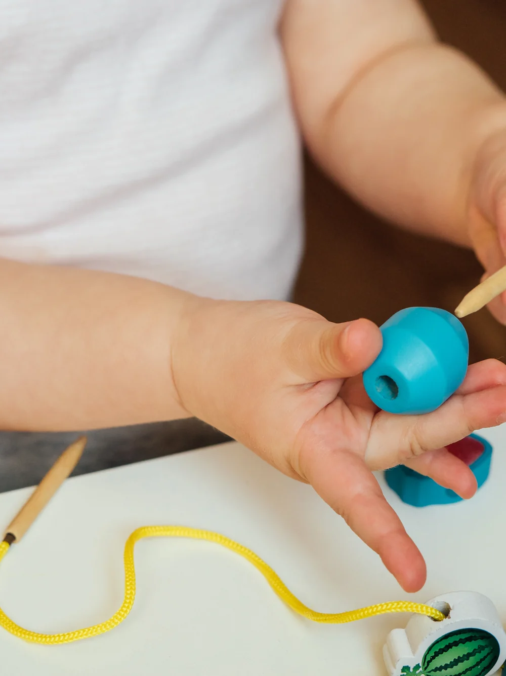 A toddler's hands thread a large blue bead onto a string at a white table