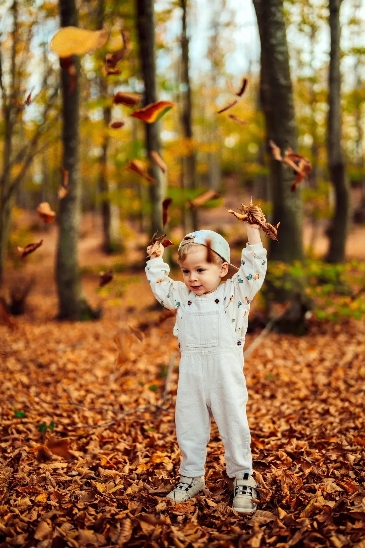 Toddler in white overalls tossing dry orange leaves into the air in an autumn woods