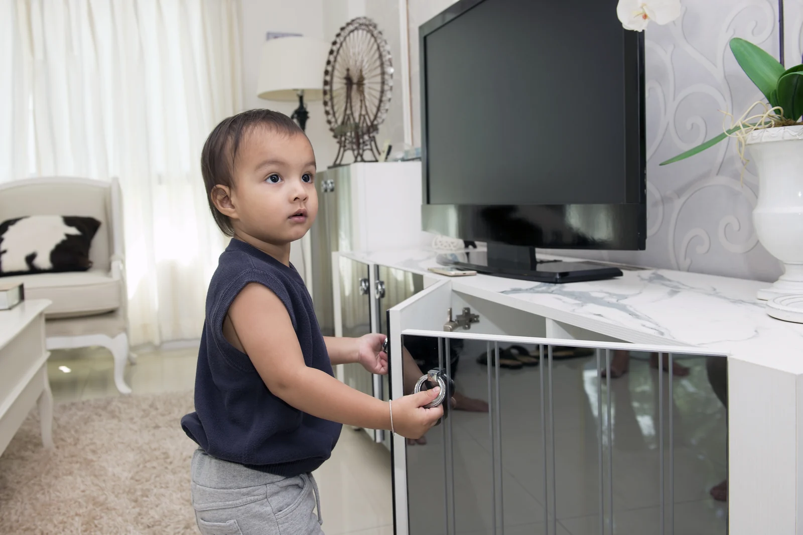 A toddler stands beside a TV stand and pulls on the ring handle of an open cabinet door