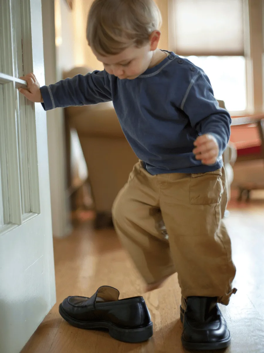 Toddler sitting on the floor by the door trying to pull on a shoe by themselves