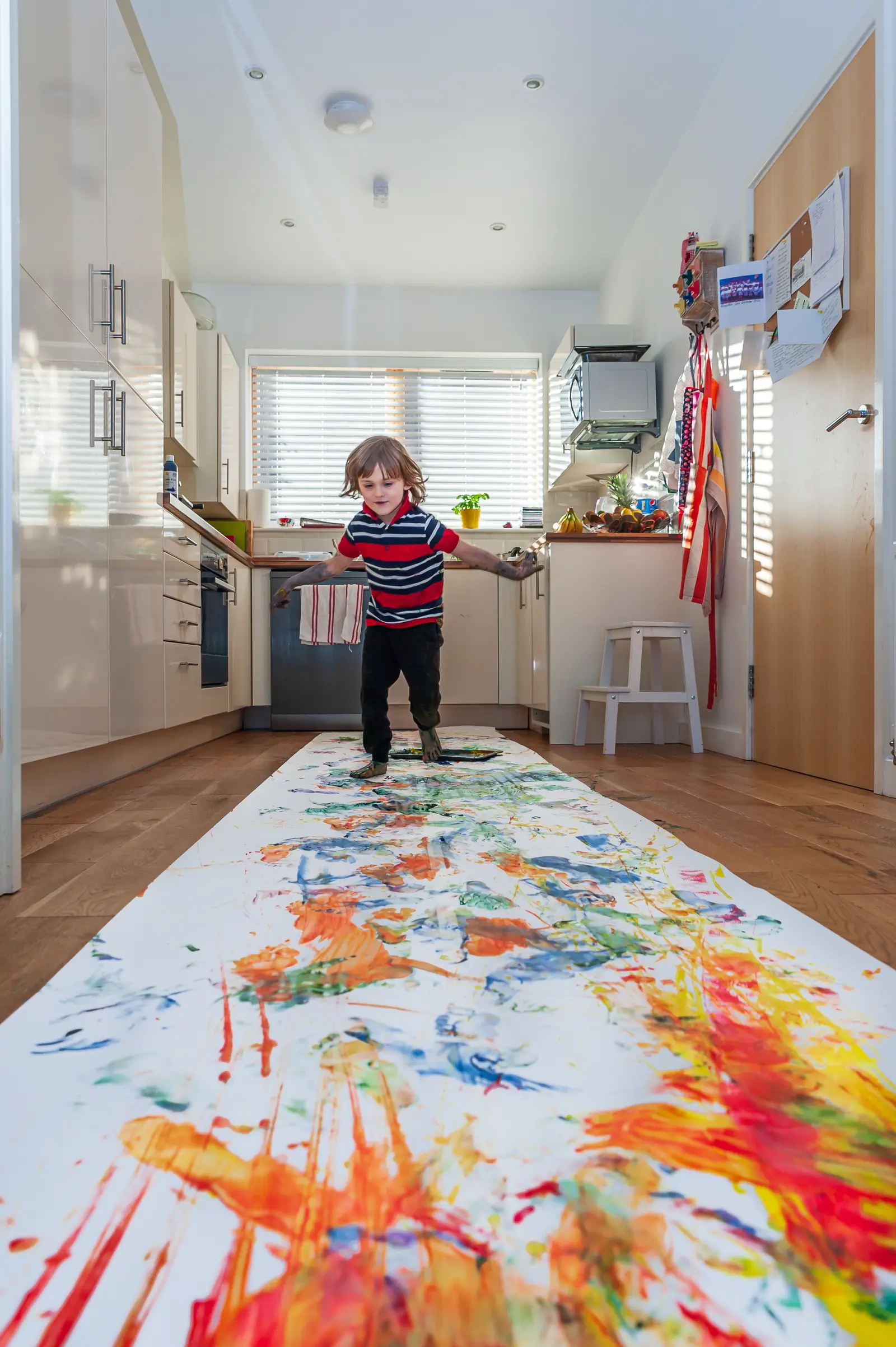 Child walking along a long strip of paper covered in bright paint, laid across a kitchen floor