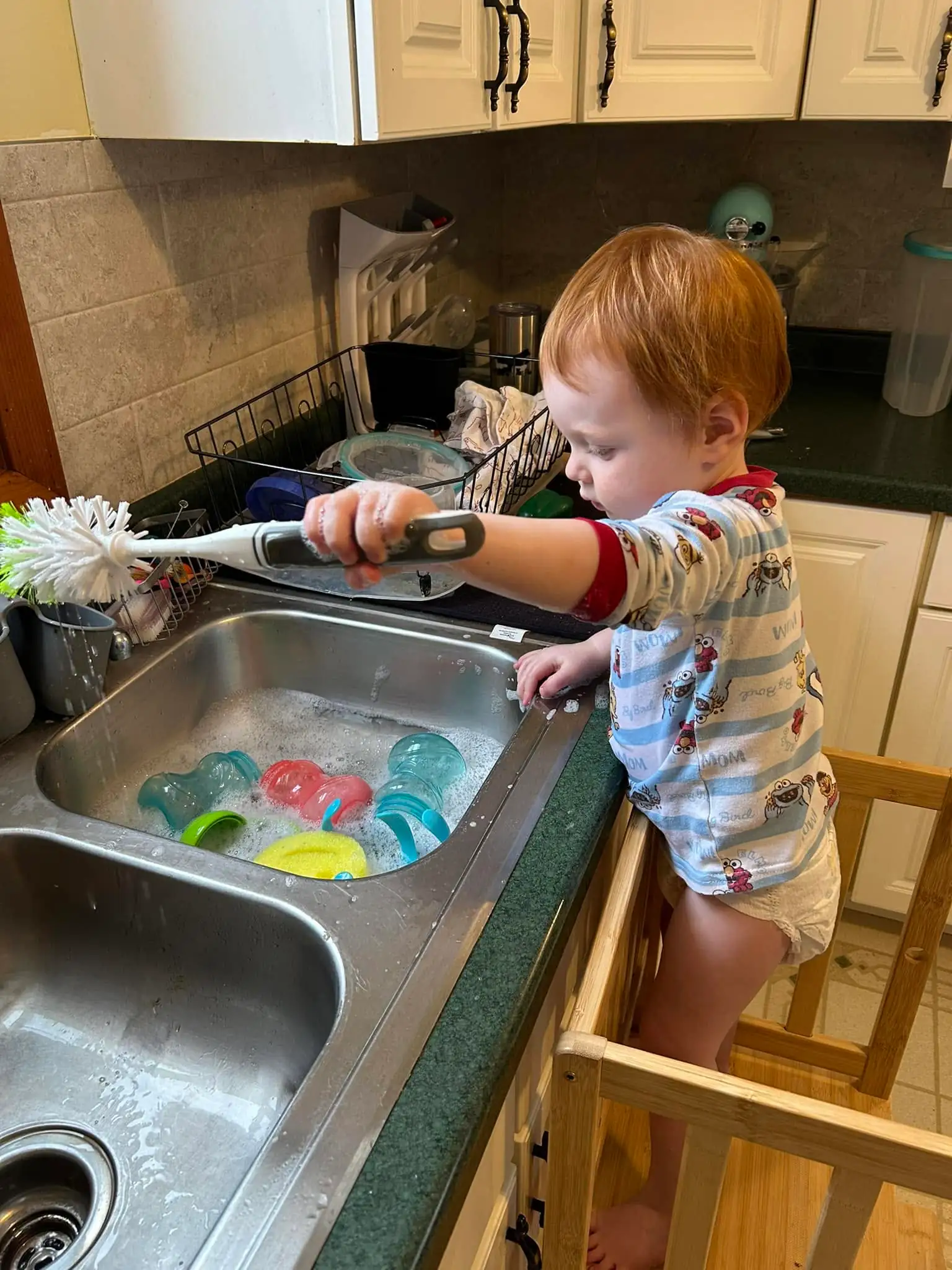 A toddler in a wooden learning tower at the kitchen sink, scrubbing cups in soapy water with a long-handled brush