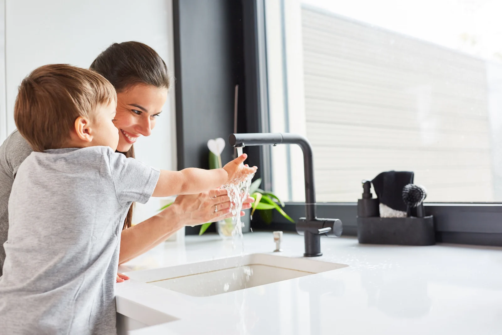 Smiling parent guiding a toddler's hands under running water at a kitchen sink