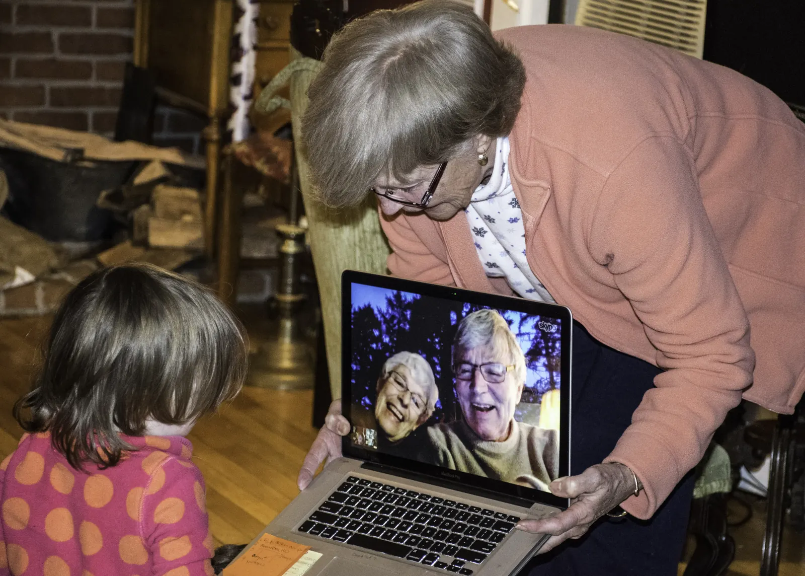 Toddler in a polka-dot top watching two smiling older people on a laptop video call held up by an older woman