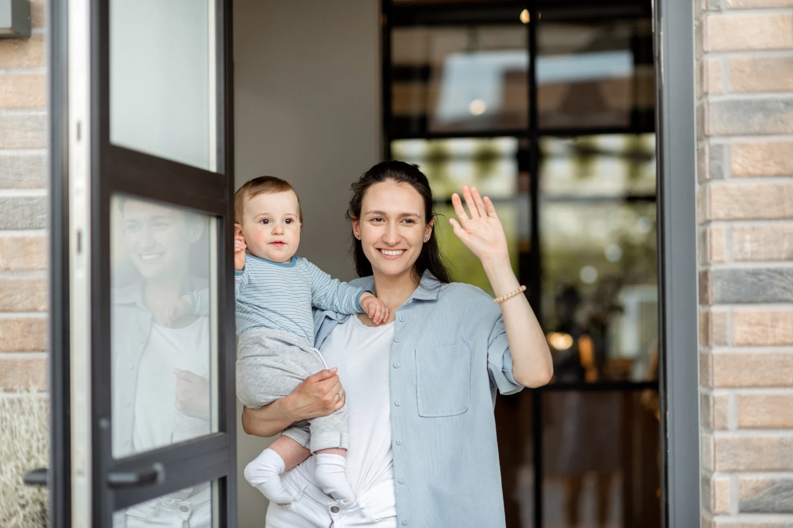 A toddler waves from an open front door while a parent stands beside them
