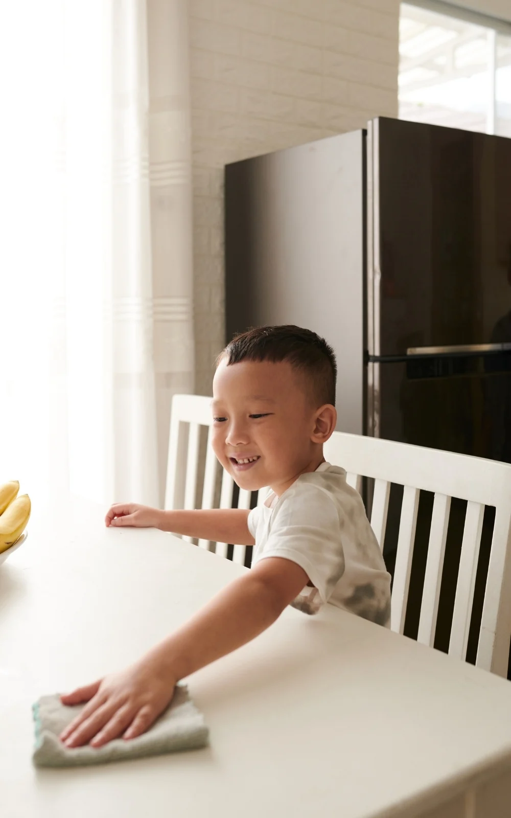 A smiling toddler leans across a white table to wipe it with a small cloth