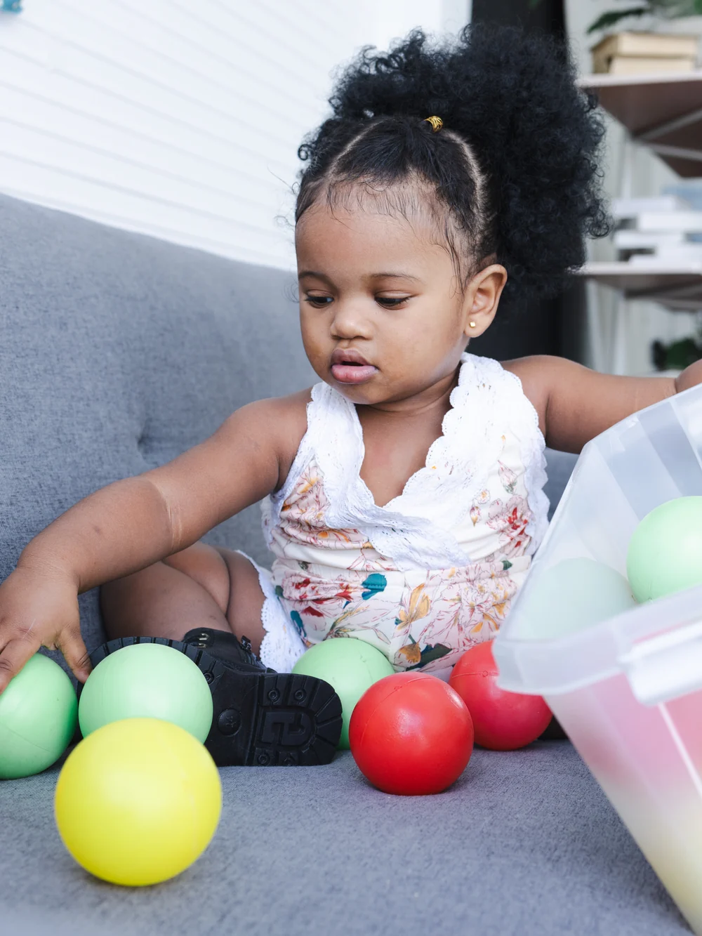 A toddler on a sofa reaches for large colorful balls beside a clear plastic tub