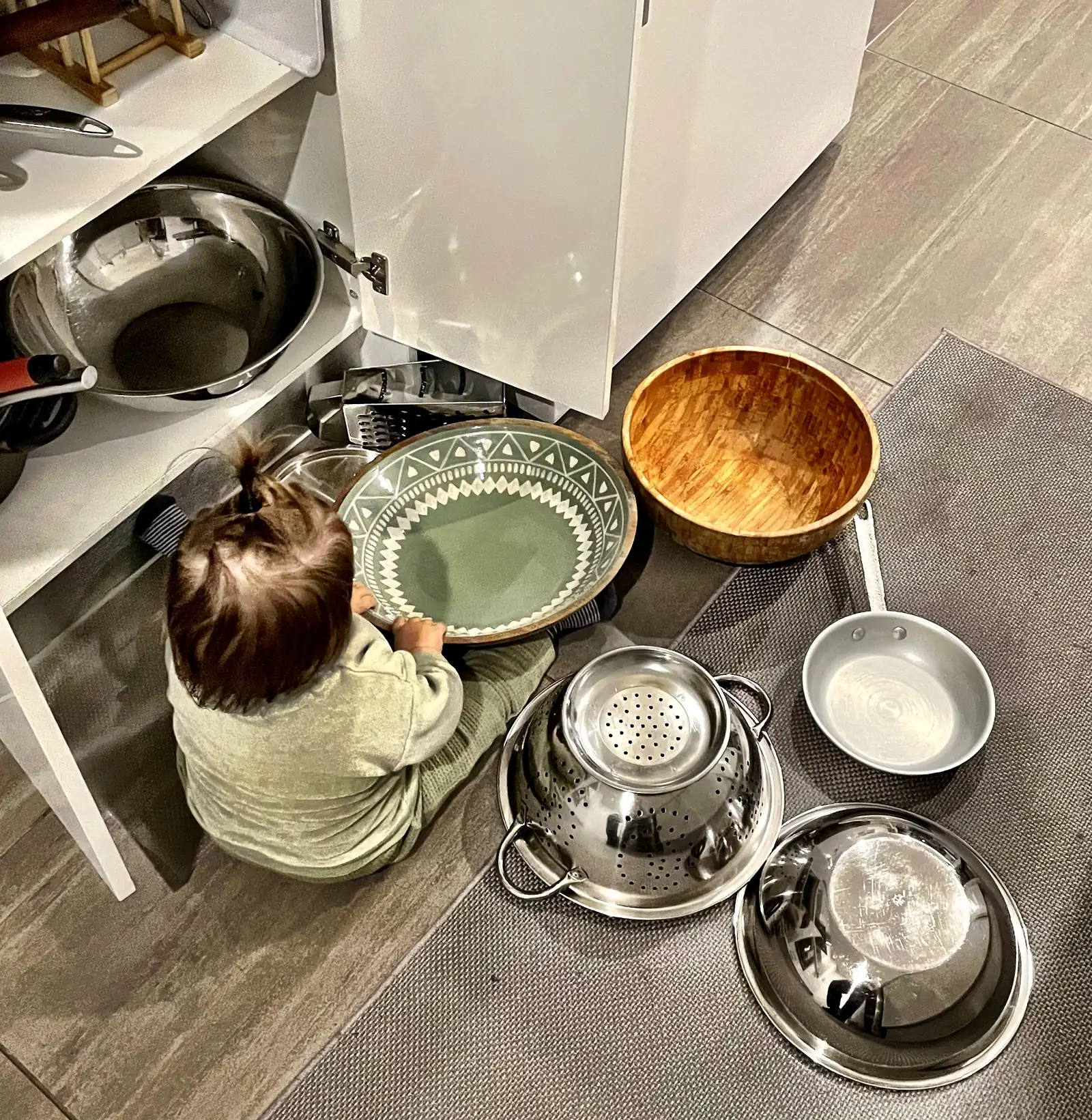 A toddler sits on a kitchen floor beside an open low cabinet, with steel bowls, a colander and a wooden bowl spread out