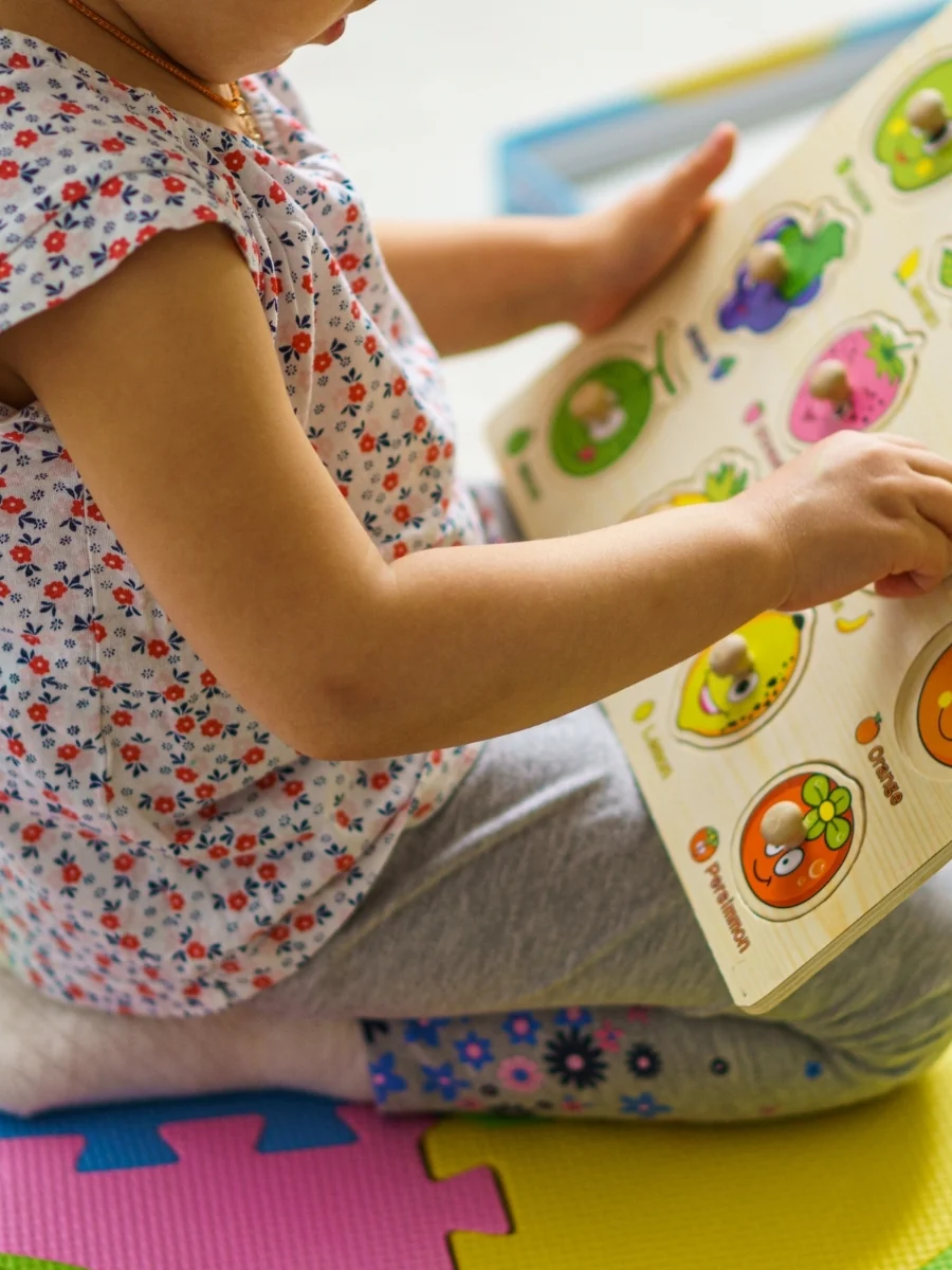 Toddler sitting on a foam play mat, holding a wooden knob puzzle with fruit shapes