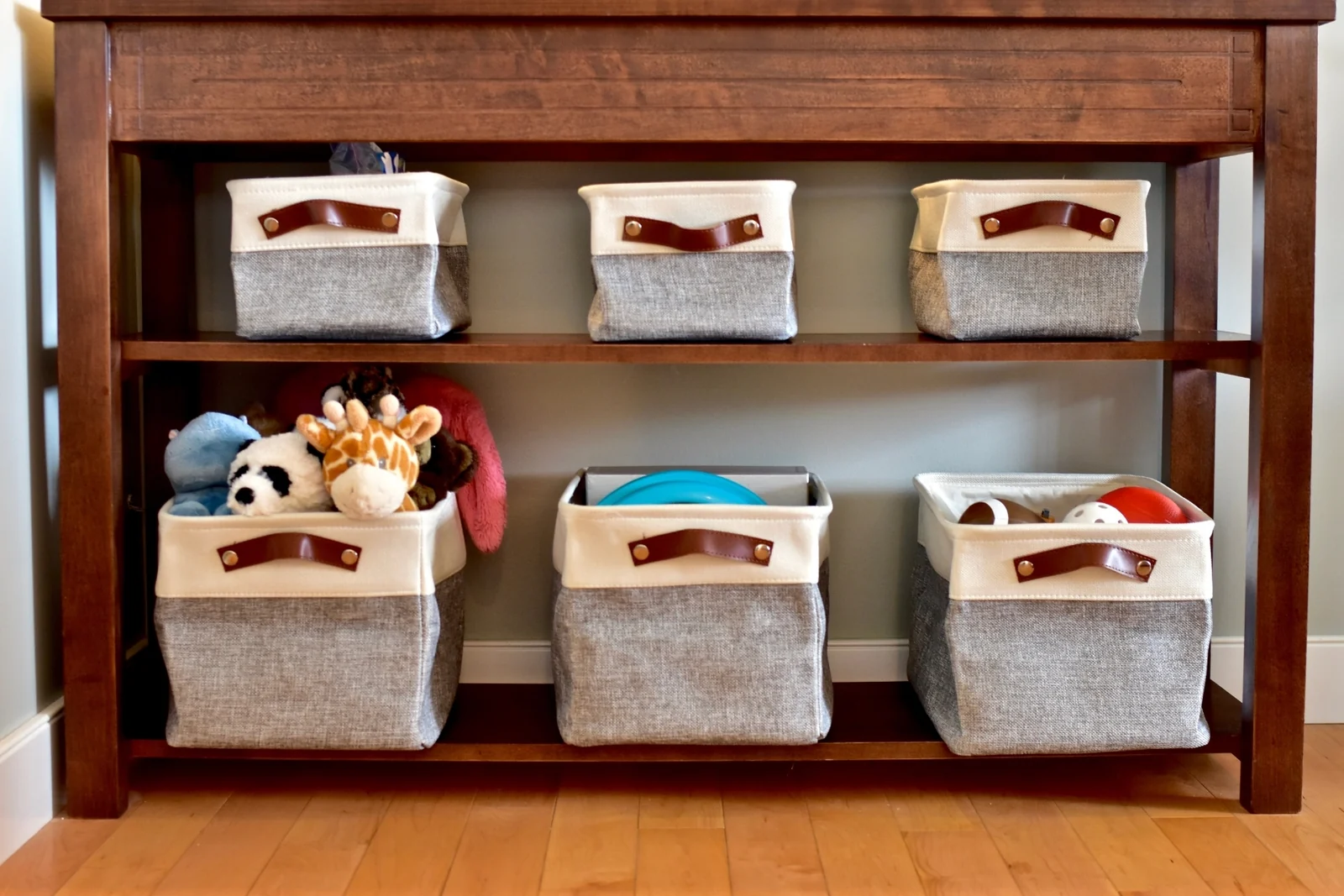 Gray and cream fabric baskets sit on two shelves of a wooden console, some holding a few toys