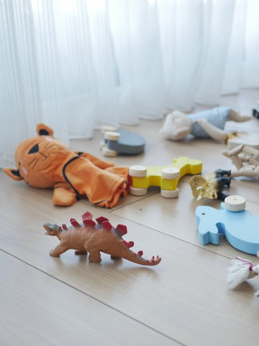 Toys scattered across a wood floor in front of a sheer curtain, including a plush crocodile, a dinosaur and wooden toys