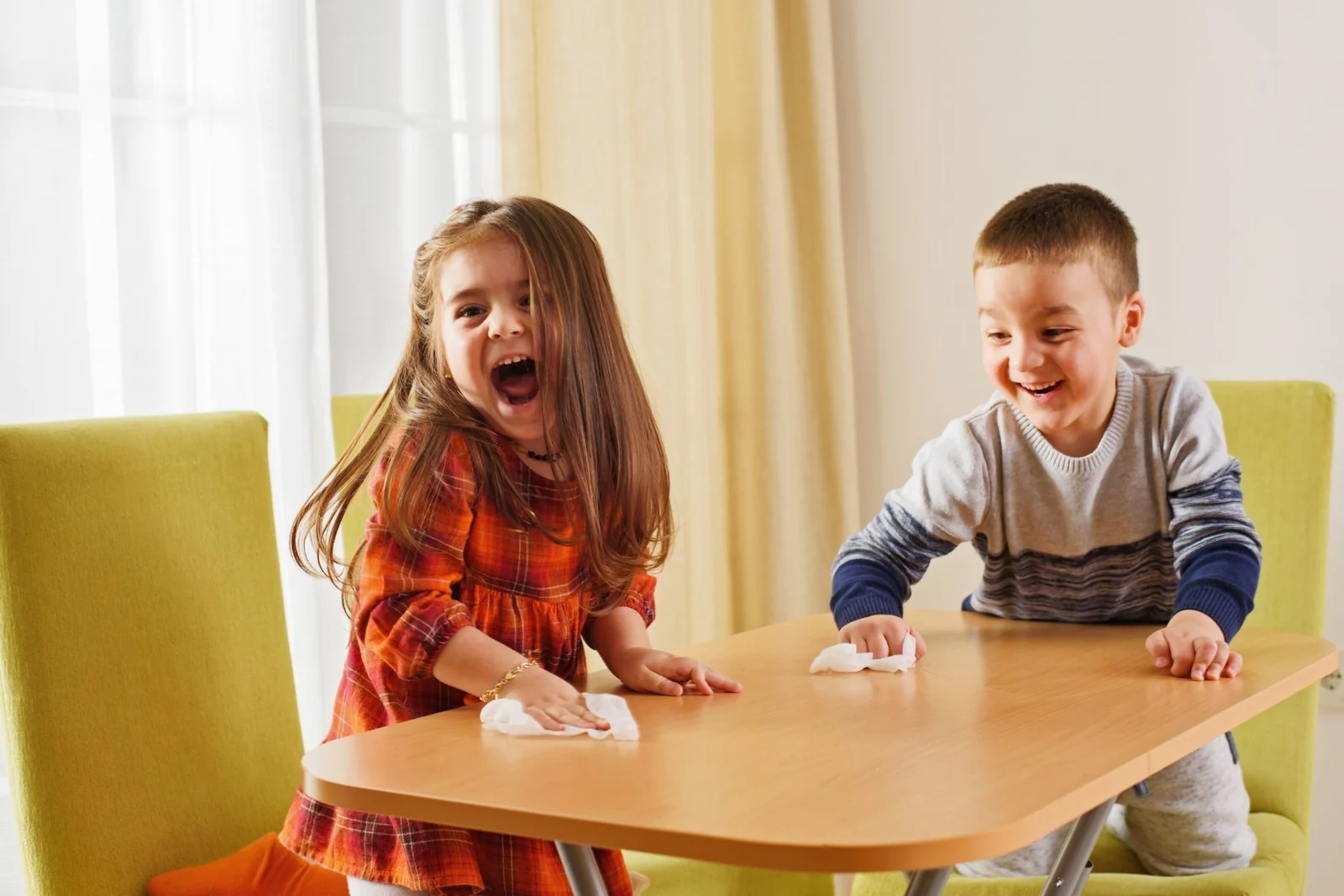 Two young children laughing as they wipe a wooden table together with small white cloths