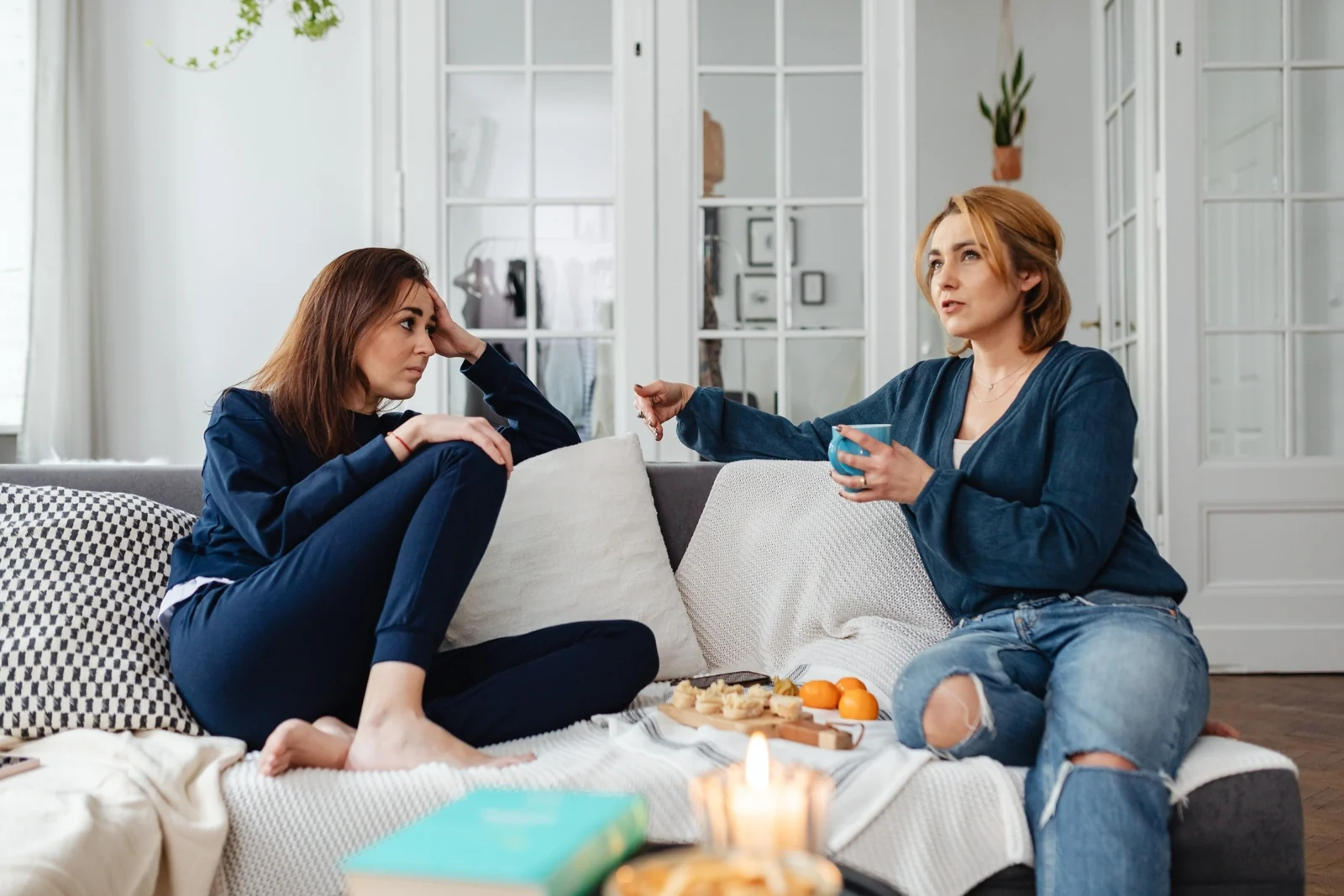 Two women sit on a sofa talking, one holding a mug, with a snack tray and candle on the table
